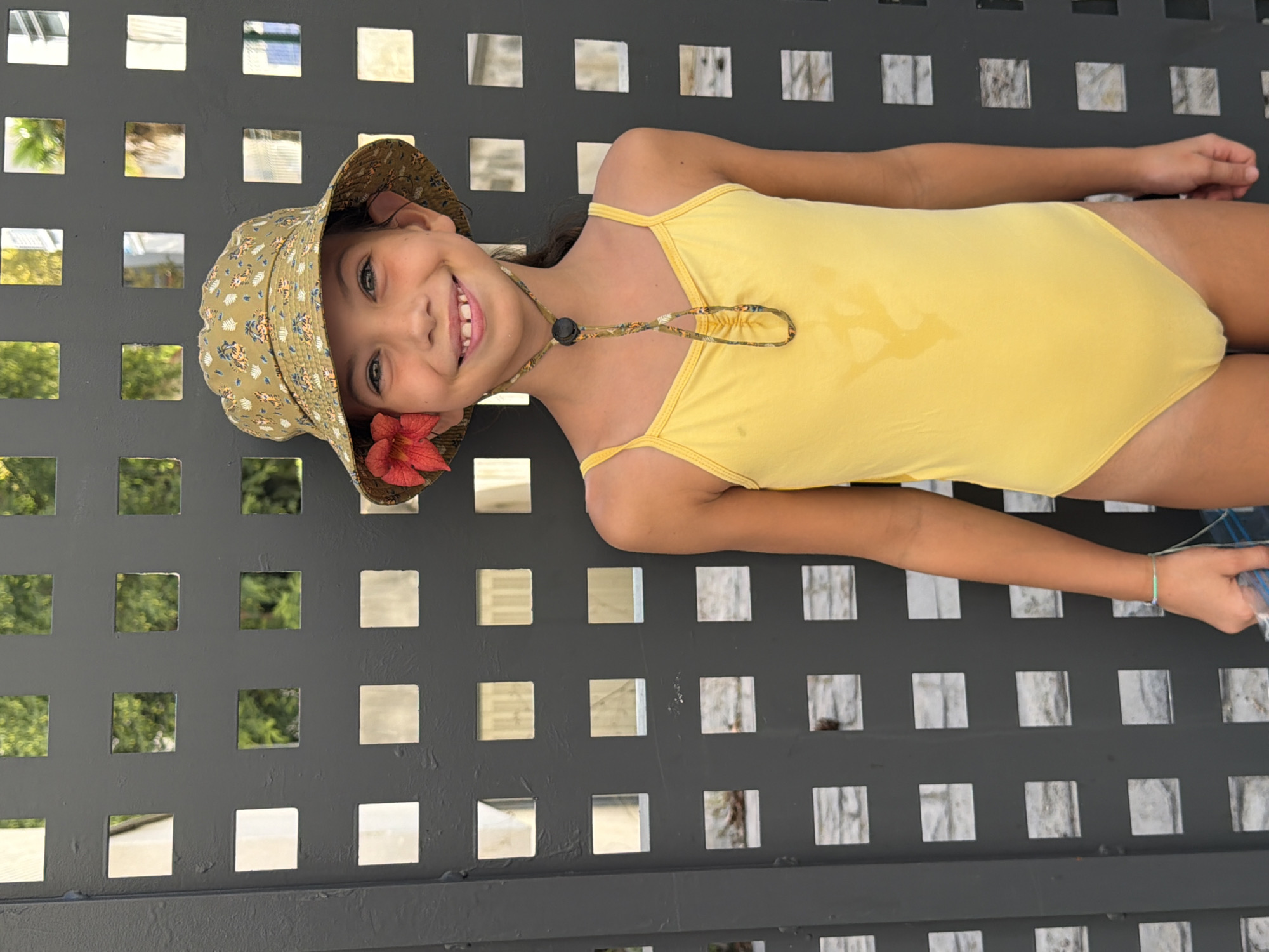 A young girl with a bright smile and a floral print bucket hat adorned with a red flower stands against a dark grey lattice fence. She is wearing a yellow one-piece swimsuit. The lattice fence has a repeating pattern of square openings, through which glimpses of green foliage and sunlight can be seen. The girl is positioned in the center of the frame, looking directly at the camera. The setting appears to be an outdoor environment, likely a residential or public space in Salou, Spain, given the sunlit foliage and architectural elements.