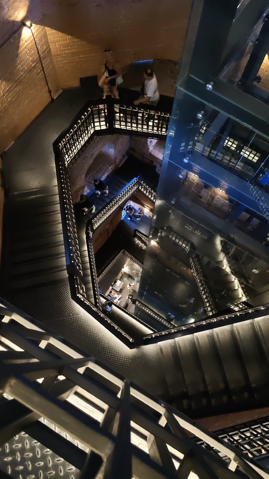 This image depicts a spiral staircase within a brick building, likely a historical structure given the brickwork and iron railings. The staircase appears to descend into a multi-level atrium or shaft. There are several people visible on different levels of the staircase, some standing and some walking. One group of people is on a landing near the top, with one person appearing to be holding a child. Another group is further down the stairs.
The lighting in the scene is dim, with artificial lights illuminating the stair treads and railings, creating a dramatic effect. The presence of glass floors or walls in the lower levels suggests a modern renovation or addition to the historical building. The reflections in the glass add to the visual complexity of the scene. The time of day appears to be evening or night due to the low ambient light and the use of artificial lighting.
The overall impression is one of architectural interest, with a blend of old and new elements. The spiral design of the staircase and the depth of the shaft create a sense of scale and perspective. The individuals on the stairs are small in comparison to the vastness of the structure, emphasizing the architectural dominance of the scene. No visible text is present in the image.
