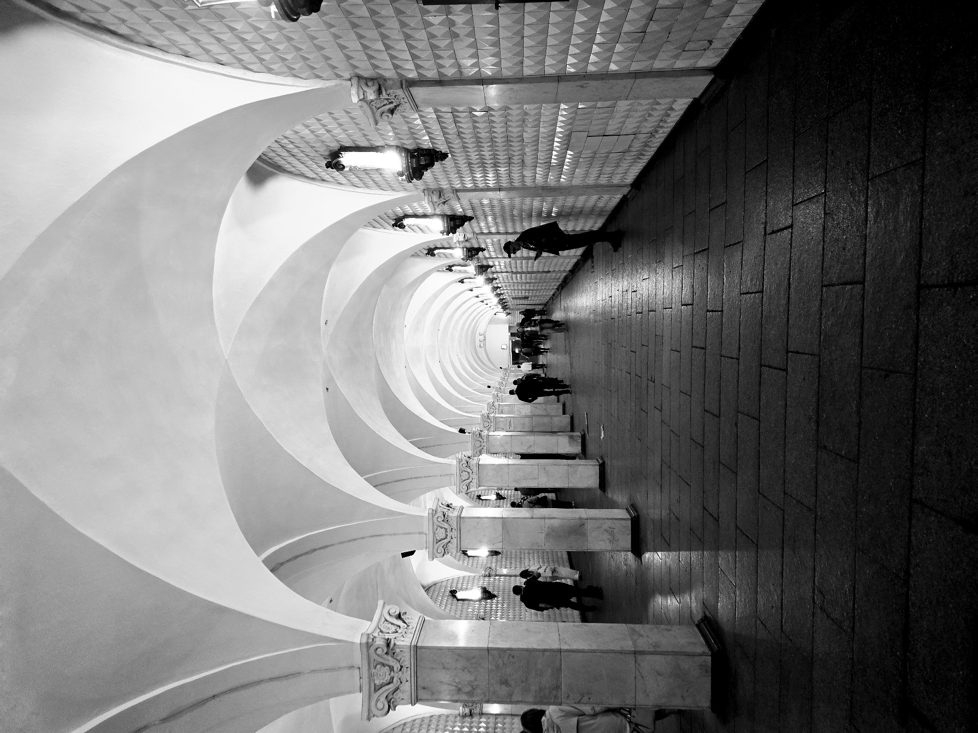 The photograph shows a black and white image of the Kiyevskaya Metro Station in Moscow, Russia. The architecture features a tunnel-like space with a series of ornate columns, white archways, and decorative lighting fixtures lining one side. The ceiling above the columns is adorned with a patterned design. People can be seen walking along the pathway next to the columns. The opposite side of the pathway is a dark wall made of rectangular tiles. The perspective of the shot emphasizes the depth and elongated shape of the tunnel.