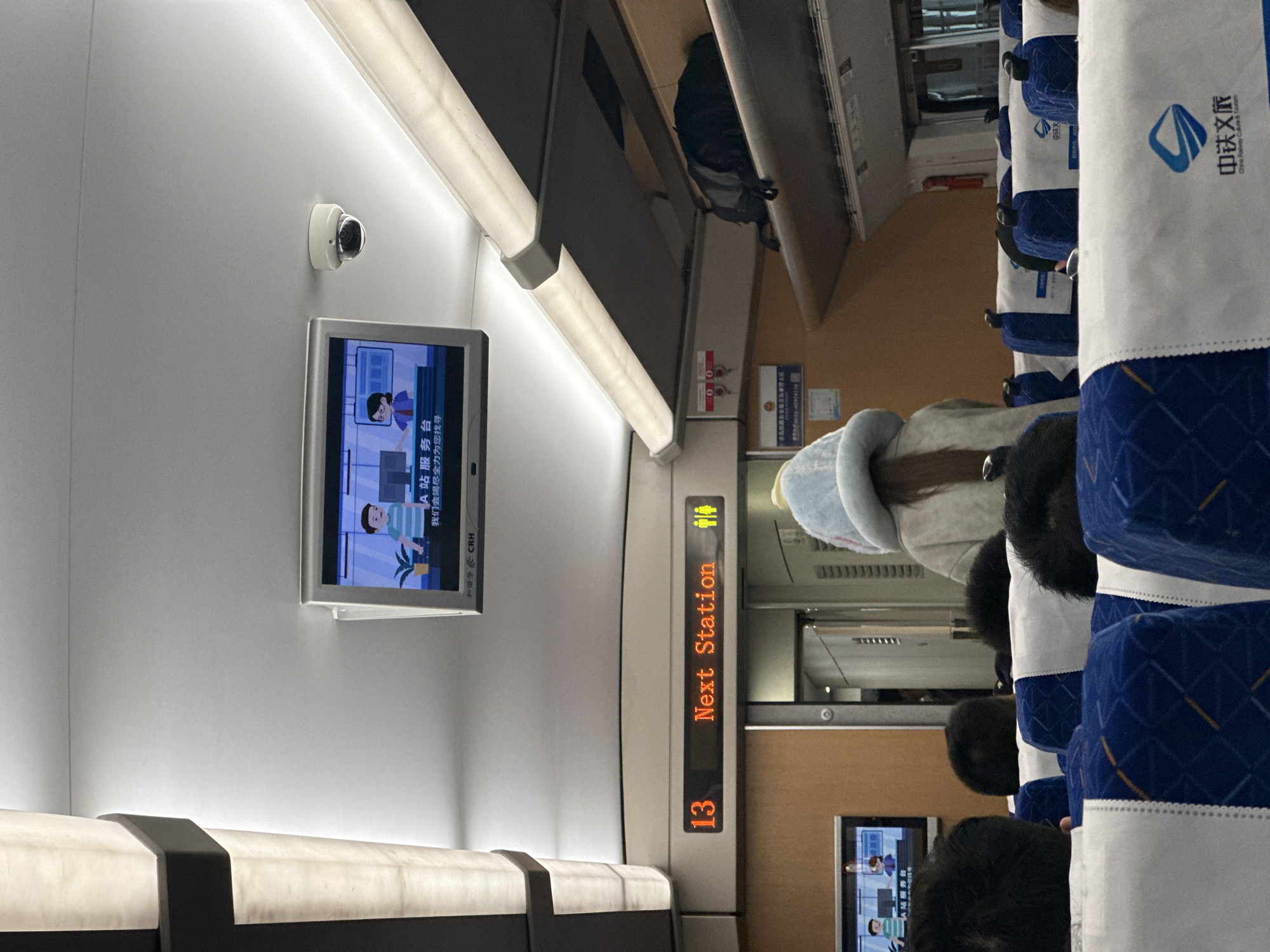 This image depicts the interior of a train carriage, identified as part of the China Railway High-speed (CRH) system, from a passenger's perspective. The location in the city is Unknown, Unknown. The scene shows multiple rows of passenger seats upholstered in blue fabric with a geometric pattern, each covered by a white headrest protector bearing the "中铁文旅" (China Railway Culture & Tourism) logo. Several passengers are visible from behind, their faces obscured. In the mid-foreground, slightly to the right of center, a passenger wears a light blue fuzzy hat with animal ears over long brown hair, paired with a light-colored top.
The ceiling is light gray/white, equipped with linear fluorescent light fixtures running along the sides. A central rectangular digital screen is mounted to the ceiling, displaying a cartoon animation featuring a male and female character in an office environment, accompanied by Chinese text and the "CRH" logo. A second, similar screen is partially visible on the upper left. A white dome-shaped CCTV camera is affixed to the ceiling near the central screen. Above the forward section of the carriage, an orange LED information display shows "13 Next Station" alongside male and female restroom pictograms. Overhead luggage racks are visible on the right, with a black backpack placed on one. A doorway leading to another part of the train is visible beyond the information display. The interior walls are a light brown or tan color. Passengers are seated, presumably in transit.