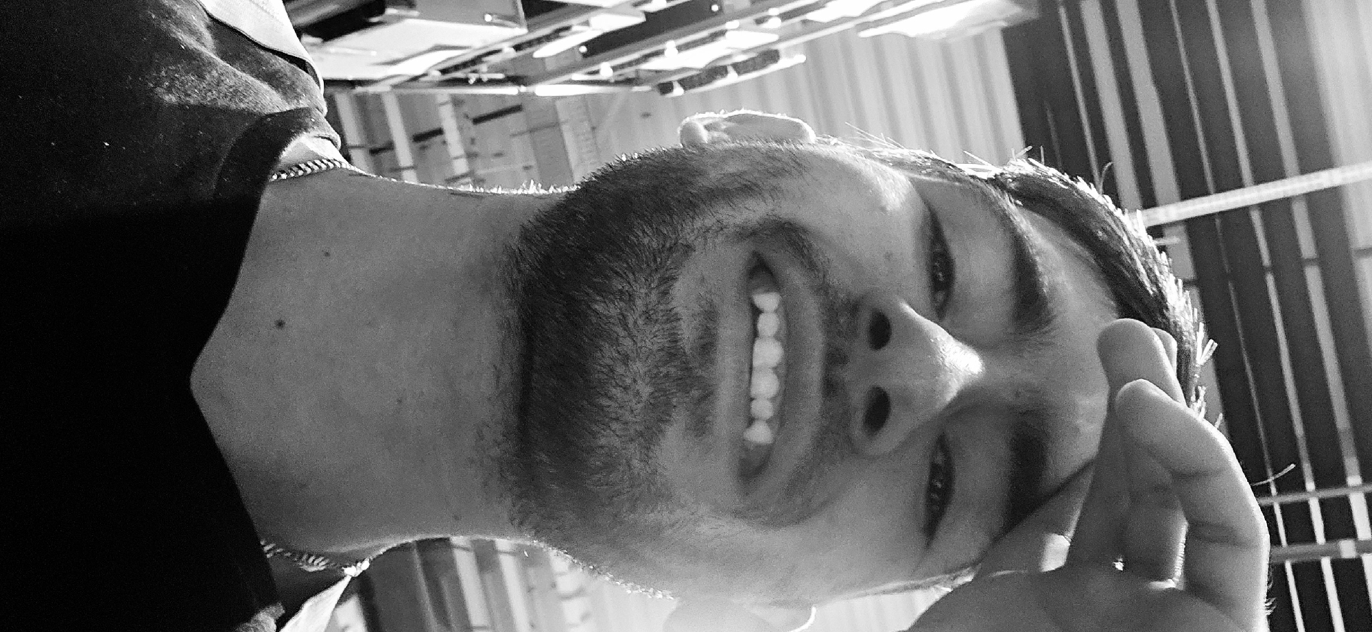 A black and white, close-up portrait features a young man smiling broadly, with his head tilted. The image is rotated, presenting the man upside down relative to a standard upright orientation. He has a short beard or prominent stubble and is wearing a dark top, possibly a t-shirt, along with a thin, metallic chain necklace visible around his neck. His wide smile reveals his upper teeth, and his eyes are partially closed in mirth. One of his hands is raised near the right side of his face, with fingers slightly curled.
The background is softly blurred but reveals a structured environment with contrasting vertical and horizontal lines and shapes, suggesting an indoor or covered outdoor space with visible architectural elements like ceiling beams or panels. The lighting appears bright, creating highlights on the man's hair and facial features, indicative of daylight or strong artificial illumination. The overall emotion conveyed is one of joy, happiness, or amusement. No visible text is present in the image. The location context provided is Plovdiv, Bulgaria.