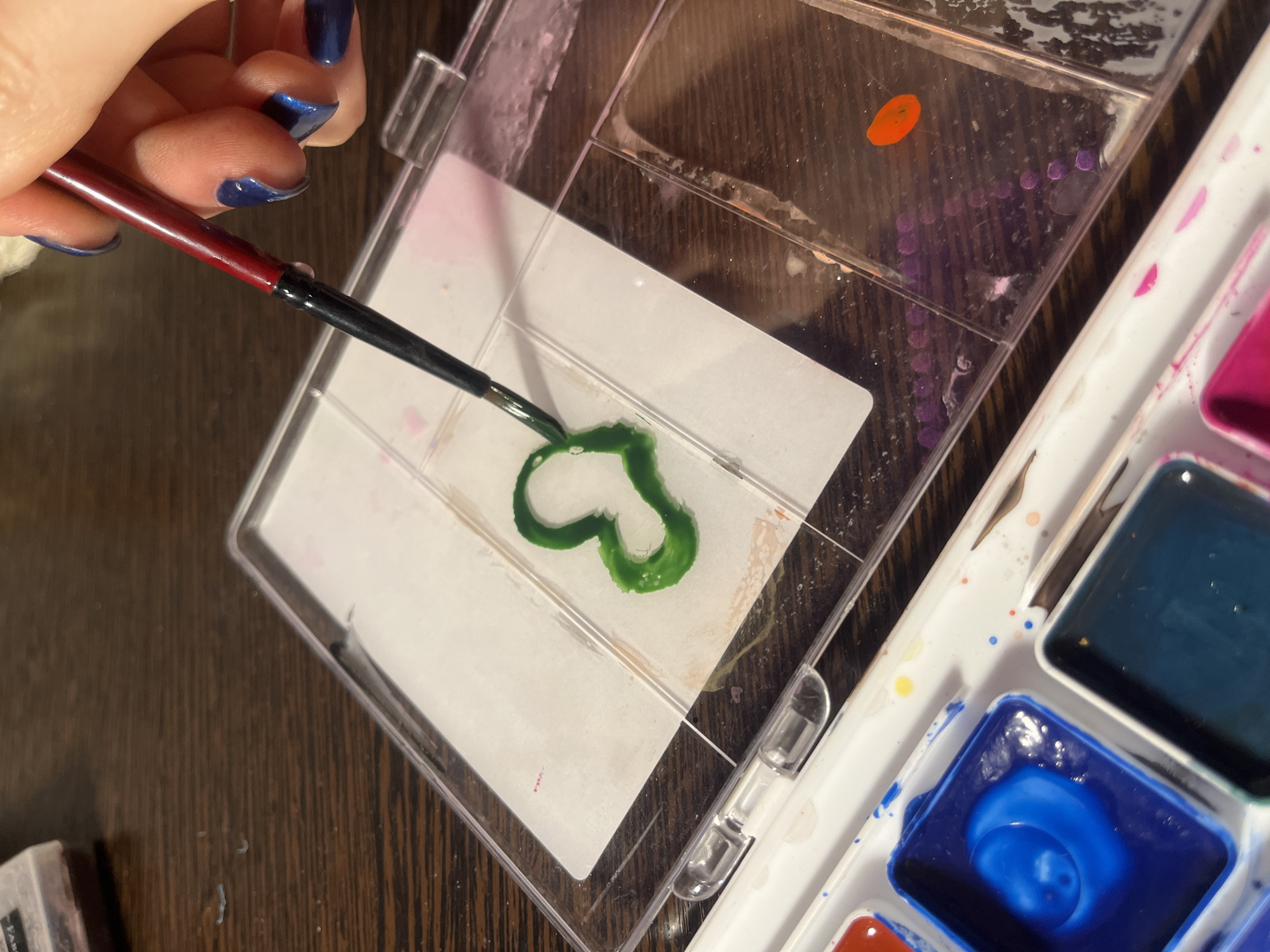 The image depicts a close-up view of a person painting with watercolors. A hand with blue nail polish holds a paintbrush with a red handle over a clear, plastic watercolor palette. On the palette, there is a blob of green watercolor paint shaped like a heart. Other colors of watercolor paint are visible in a white palette tray to the right. The background is a dark brown wooden surface. The location is unknown and not necessarily in Saint Petersburg, Russia.