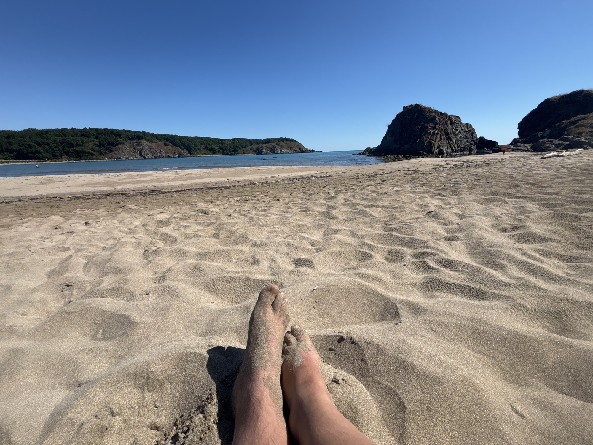 The image depicts a beach scene with a first-person perspective of a person's sandy feet in the foreground. The sand is rippled, indicating wind action. In the mid-ground, a calm body of water is visible, meeting a sandy shoreline. Beyond the water, lush green hills with some rocky outcrops form the background, under a clear, bright blue sky. The location is identified as a beach in Rezovo, Bulgaria.
