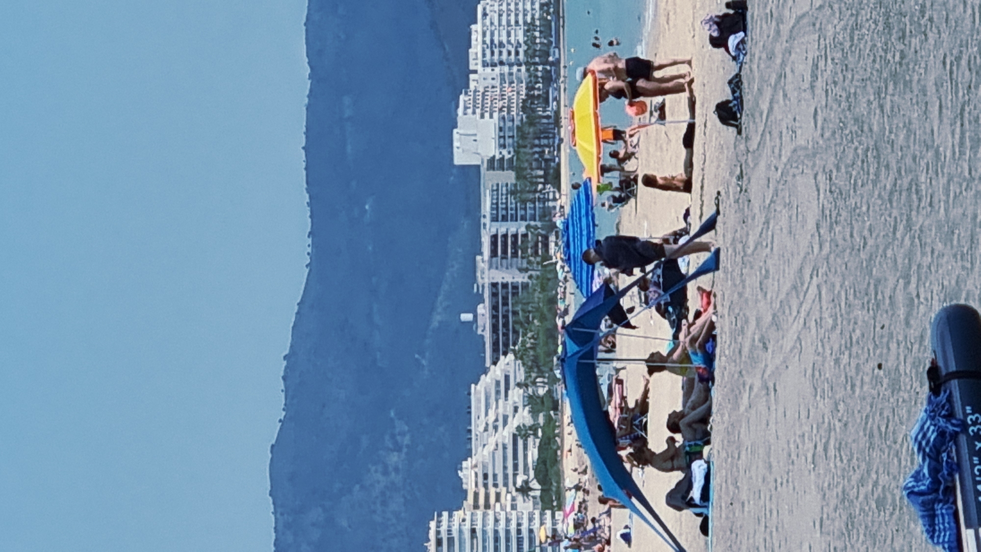 The image depicts a crowded beach scene with several people lounging under umbrellas. In the foreground, a large blue umbrella shades a group of individuals. To the right, a yellow umbrella shelters a couple. Further back, numerous other beachgoers and umbrellas are visible. Beyond the beach, a series of modern, multi-story buildings line the coast, suggesting an urban or resort setting. In the distance, a range of blue-hued mountains forms the backdrop. The overall environment is sunny and appears to be a typical summer day at the beach. The presence of buildings and the general layout of the scene are consistent with the coastal areas of Castelló d'Empúries, Spain, a municipality known for its beaches and tourist infrastructure.