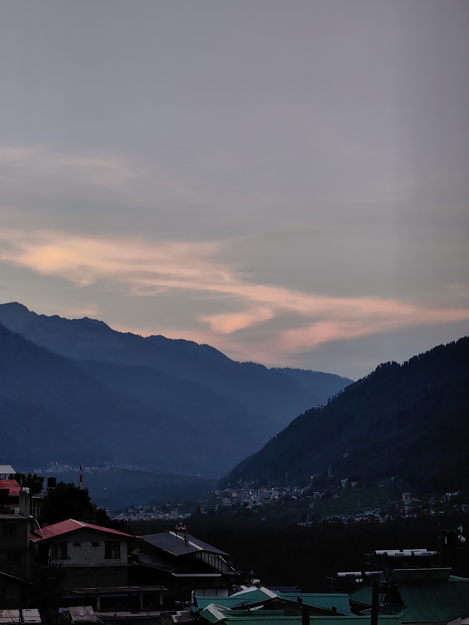 The image depicts a valley scene at dusk in Vashisht, India, with layered mountain ranges fading into the distance. The sky is a soft gradient of pale blue and grey, streaked with wispy clouds tinged with pale pink from the setting sun. Dark, silhouetted mountains dominate the middle ground, with lighter shades indicating receding peaks. In the foreground, a cluster of buildings with red and dark-colored roofs are visible, nestled against the foothills. A town or village is spread across the valley floor, its lights faintly twinkling in the dimming light. The overall atmosphere is serene and twilight.