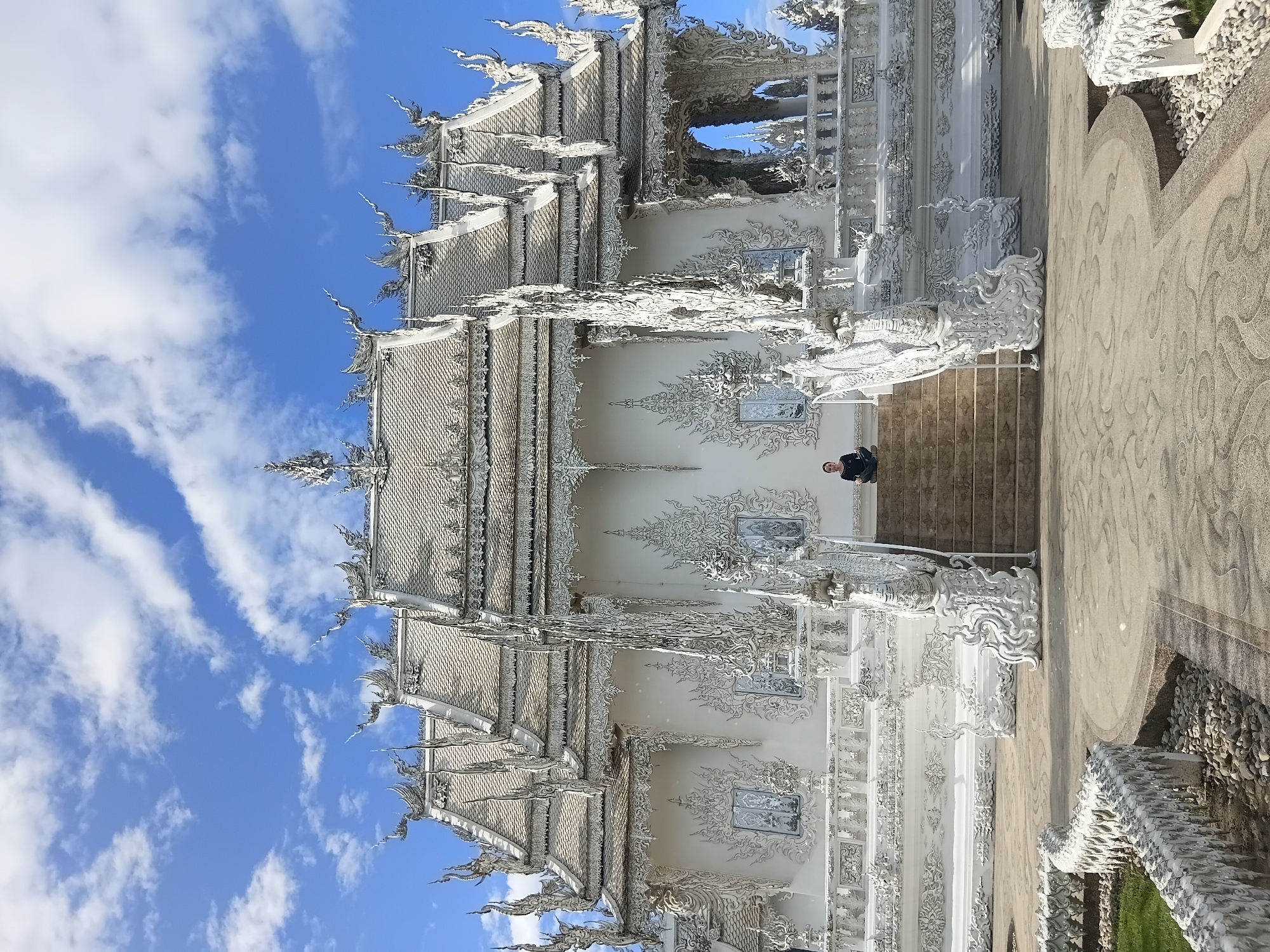 A person is sitting cross-legged on the middle of a set of stairs leading up to a white building, with their eyes closed. The building is highly ornate with intricate white decorations on a white facade. The sky above is blue with scattered white clouds. The location is the Wat Rong Khun, also known as the White Temple, in Ban Rong Khun, Thailand.