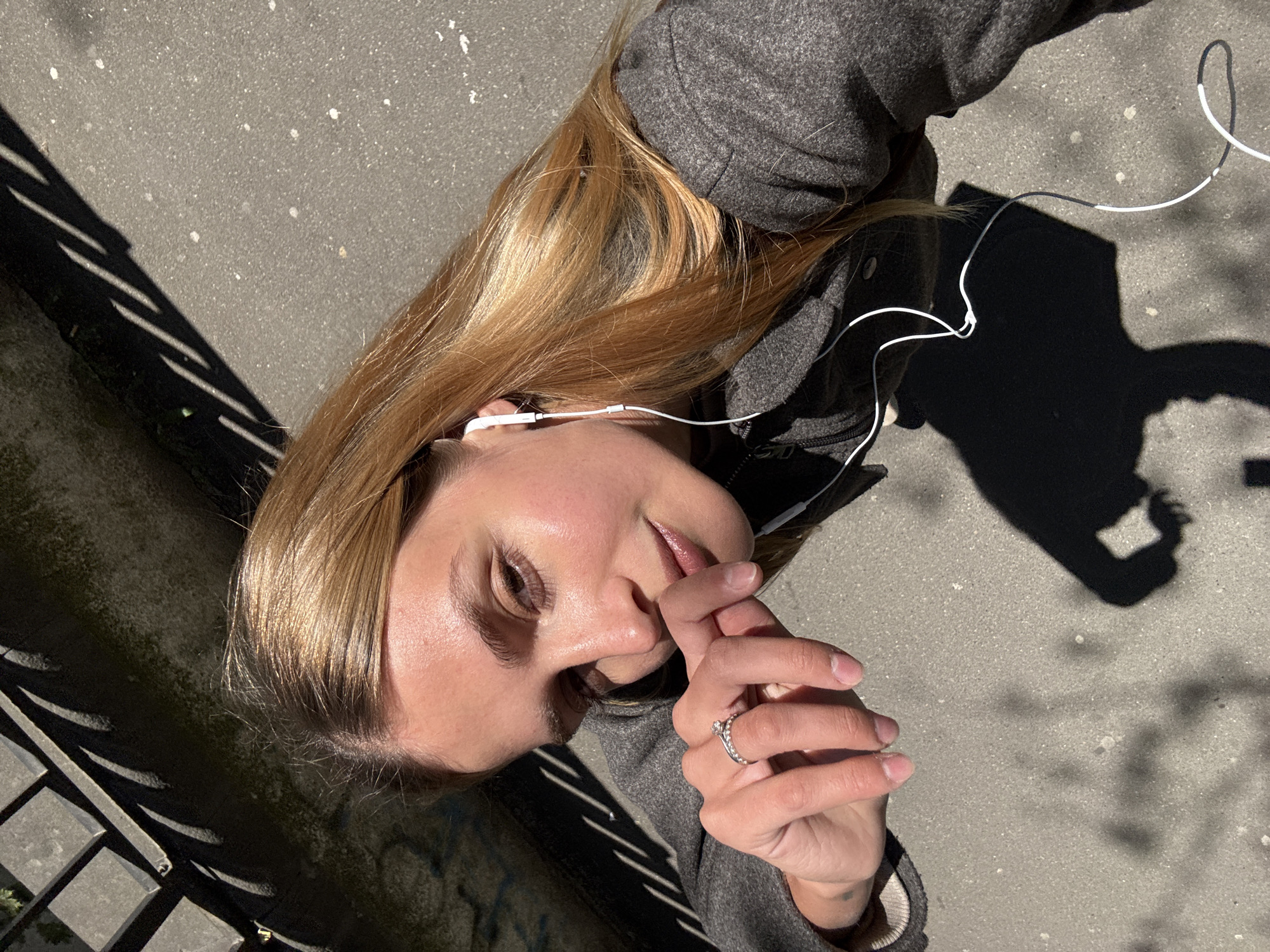 A woman lies on a paved surface, possibly a sidewalk, in Tbilisi, Georgia. She has fair skin and long blonde hair. She wears a gray coat and white earbuds. Her right hand is raised near her face, with a ring visible on her finger. The pavement is cracked and dirty. There are shadows from a metal fence on the left and an unidentified object to the right.