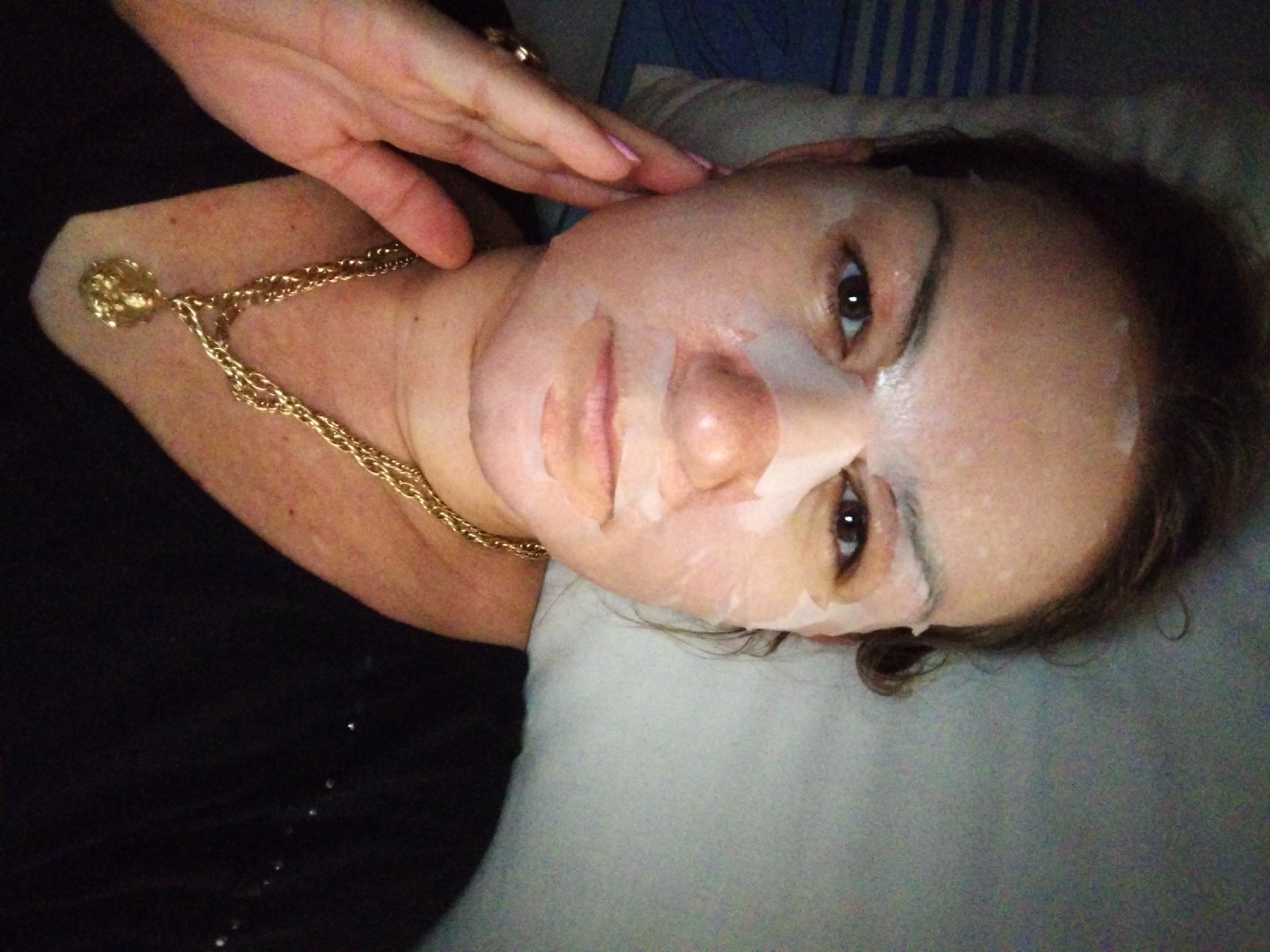 A female subject is depicted indoors in Stara Zagora, Bulgaria. She is captured in a close-up, chest-up shot, looking directly at the camera. Her face is covered with a translucent white sheet mask, which adheres to her skin, leaving her eyes, nostrils, and mouth exposed. Her left hand, wearing a gold ring on the ring finger and exhibiting pink nail polish on the visible fingers, is gently resting against her left cheek and jawline. She has dark hair, brown eyes, and is wearing a dark, possibly black, long-sleeved garment with subtle shimmering details on the right shoulder. Around her neck is a multi-strand gold chain necklace with a circular, intricately detailed gold pendant. The background on the left side features a blue and white striped pattern, consistent with bedding or a pillow. The background on the right is a plain, light-colored wall. The scene is dimly lit, suggesting an indoor setting during evening or night hours.