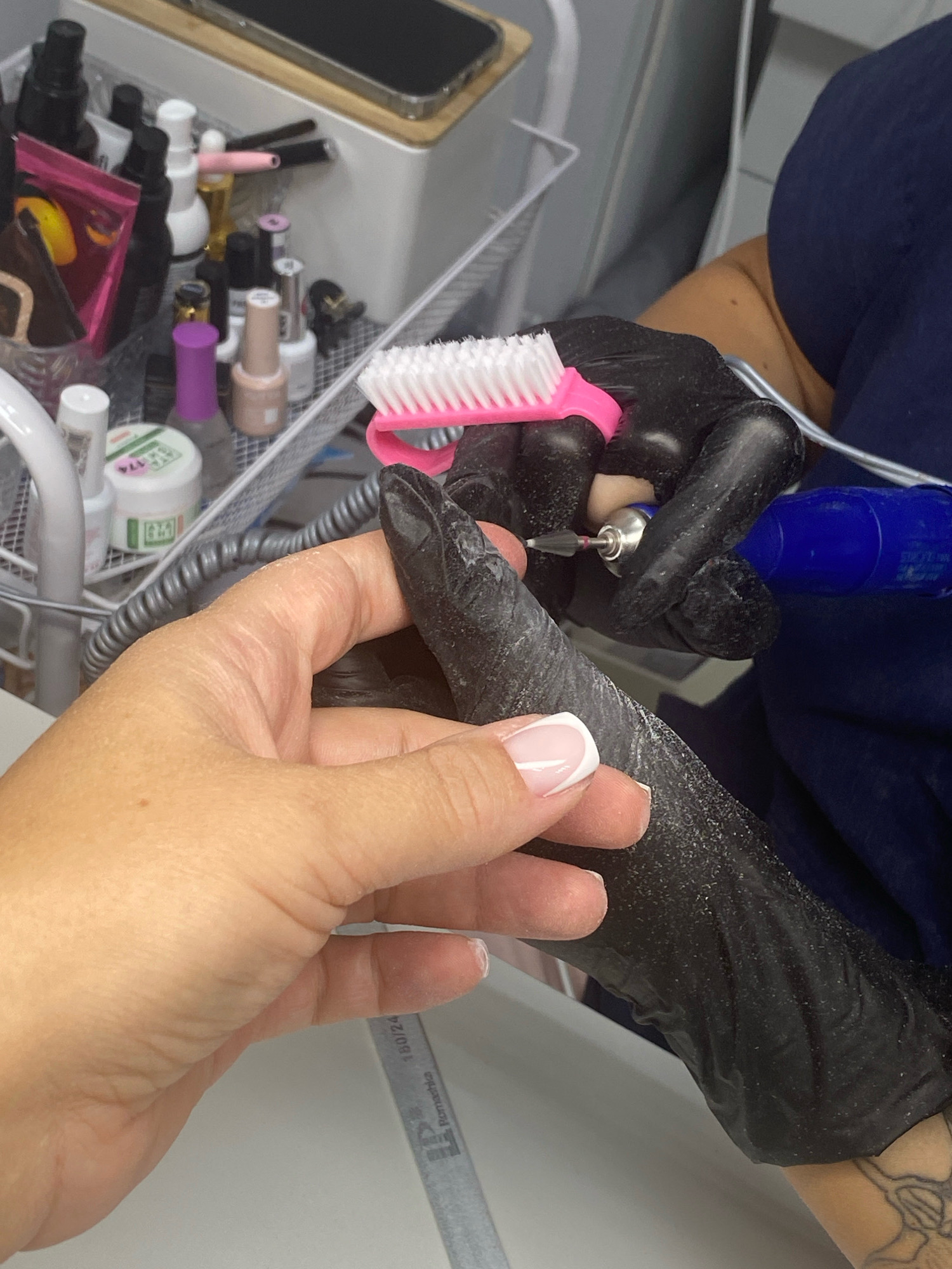 This is a close-up, eye-level, medium shot of two hands with black gloves on one of them, performing a nail service. The hand on the right, wearing a black glove, is holding an electric nail drill and a small pink brush with white bristles. The drill bit is actively working on the nail of the hand on the left. The hand on the left is bare and shows a well-maintained nail with a French manicure. The background is blurred but suggests a salon environment with various nail care products on a tiered cart. The lighting is bright, illuminating the hands and the task at hand. This scene is not specific to Sochi, Russia; it is a general depiction of a nail salon service.