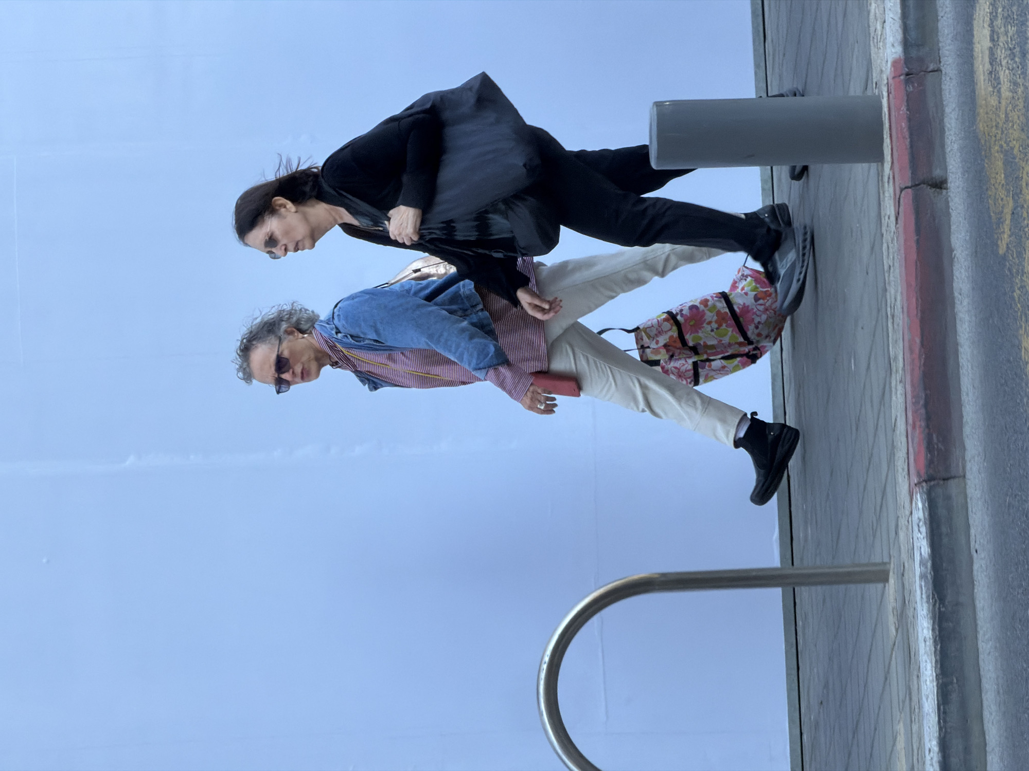The photograph shows two individuals walking on a sidewalk in Tel Aviv, Israel. The first person is a woman wearing sunglasses, a black blouse, and black pants, with a large grey tote bag over her shoulder. The second person appears to be a man with sunglasses, a red and white striped shirt, and a denim jacket, also carrying a floral-patterned tote bag and holding a red phone in his hand. They are walking on a paved sidewalk next to a grey bollard and a metal railing. A section of the curb is visible, with traces of red paint along the side. The backdrop is predominantly the sky.