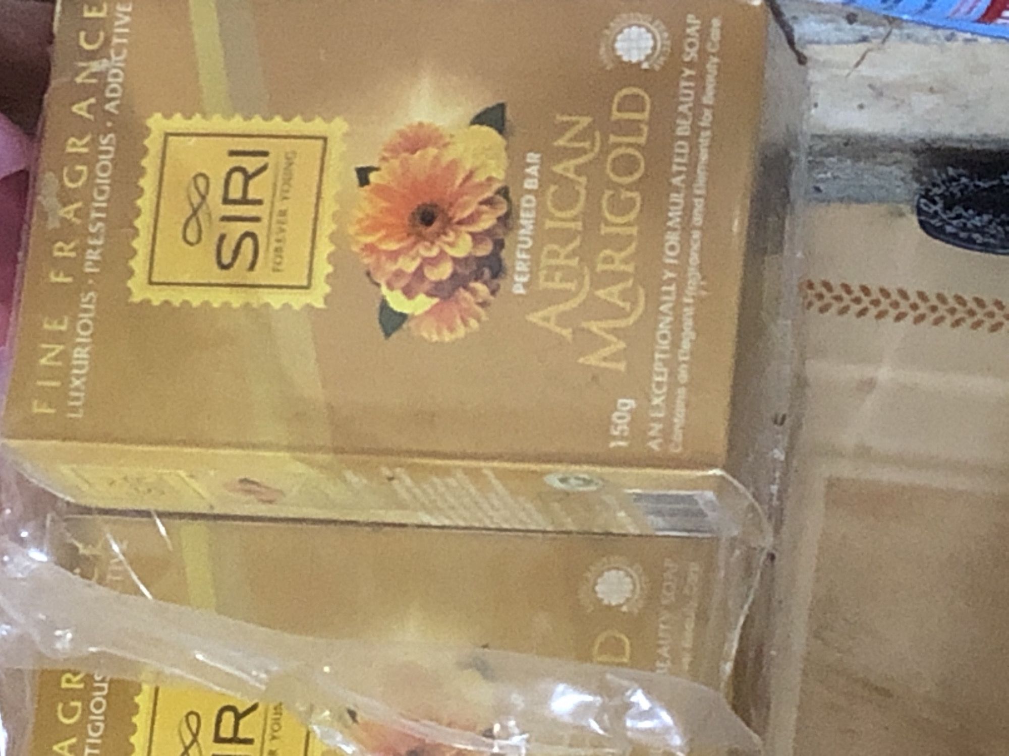 The image displays two stacked boxes of what appears to be soap. The top box is mostly obscured by a plastic wrapper and only the bottom portion is visible, showing text that includes "BEAUTY SOAP" and a circular emblem. The bottom box is more clearly visible and features an "AFRICAN MARIGOLD PERFUMED BAR" design with an illustration of marigold flowers. It also includes text that reads "150g" and "EXCEPTIIONALLY FORMULATED BEAUTY SOAP". Towards the right side of the bottom box, there is a yellow stamp-like logo with the word "SIRI" and "FOREVER YOUNG" printed vertically. The boxes are gold in color and are arranged in a retail setting, likely on a shelf. There are no people or explicit indications of location within the city of Kabong, Nigeria, visible in the image.