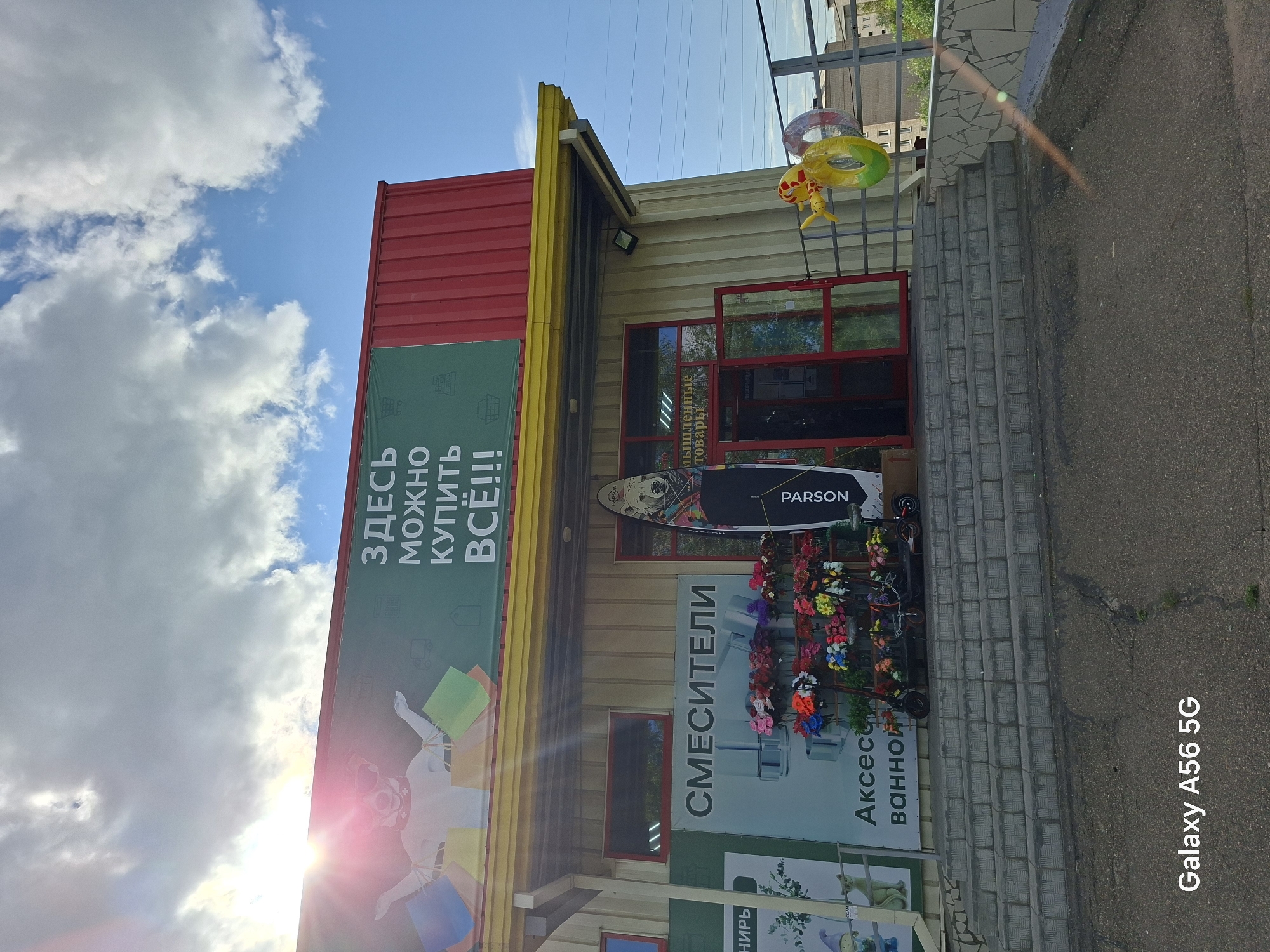 The image depicts the exterior of a store in Chita, Russia, on a sunny day with clouds. A large red and yellow sign above the entrance reads "ЗДЕСЬ МОЖНО КУПИТЬ ВСЁ!!!" (Here you can buy everything!!!). Below the sign, a billboard displays cartoon images and text. A surfboard with a polar bear design stands upright near the entrance. Various items are displayed outside the store, including what appears to be inflatable rings and a collection of artificial flowers. In front of the store, there are steps leading up to the entrance. The store's facade is made of light-colored metal siding. The ground in front of the steps is paved with asphalt. The text "Galaxy A56 5G" is overlaid on the bottom left of the image.