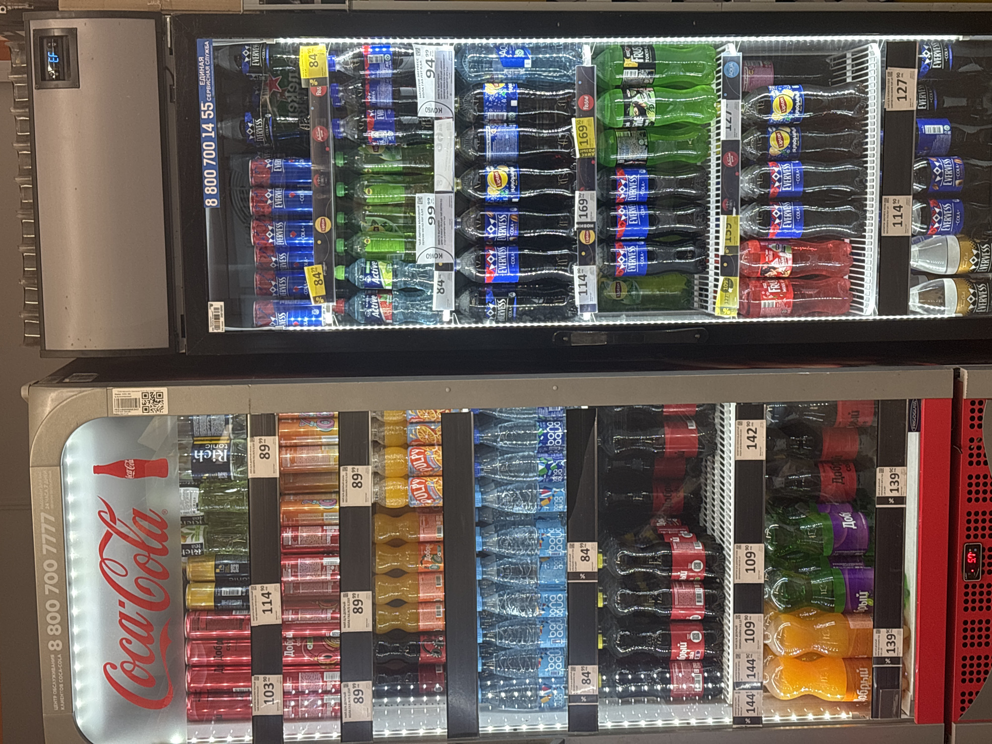 The image shows a brightly lit commercial refrigerator filled with a variety of bottled and canned beverages. The refrigerator is divided into multiple shelves, each stocked with different brands and types of drinks, including sodas, juices, and water. Labels and price tags are visible on many of the products. The refrigerator appears to be in a retail setting, possibly a convenience store or supermarket. The overall condition of the refrigerator and its contents suggests a well-maintained display. The image was taken in Sinyavino, Russia.