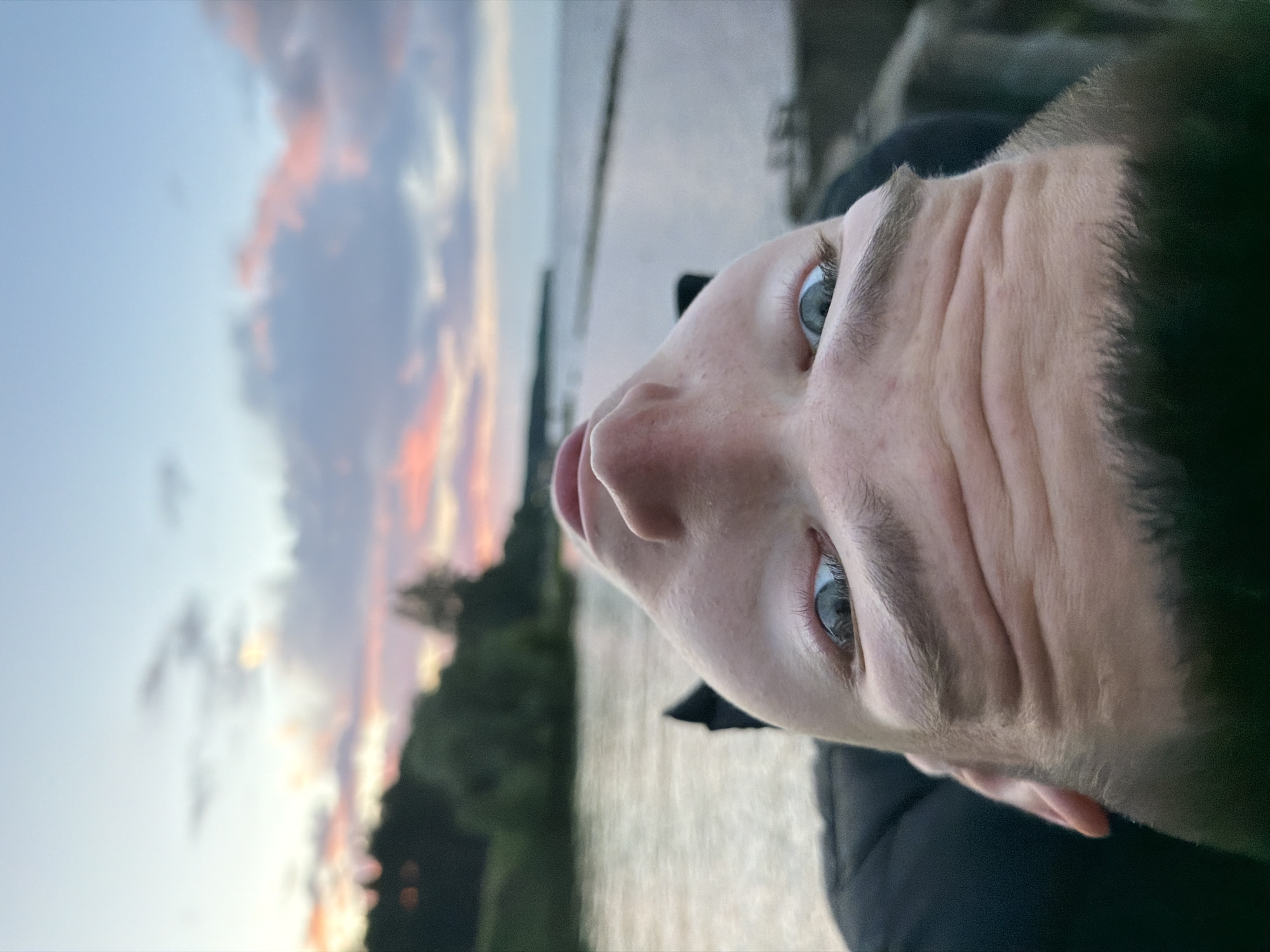An extreme close-up, upside-down selfie captures the face of a young Caucasian person looking upwards at the sky. The person has light skin, light blue eyes with visible eyelashes, and thick eyebrows. Their face is creased, indicating an upward gaze or a smile. They are wearing a black jacket. The background shows a body of water with a distant shoreline lined with trees, likely the Kama River in Chaikovsky, Russia, under a sky with dramatic sunset clouds painted in hues of pink, purple, and blue. The composition is distorted due to the unusual angle of the shot.