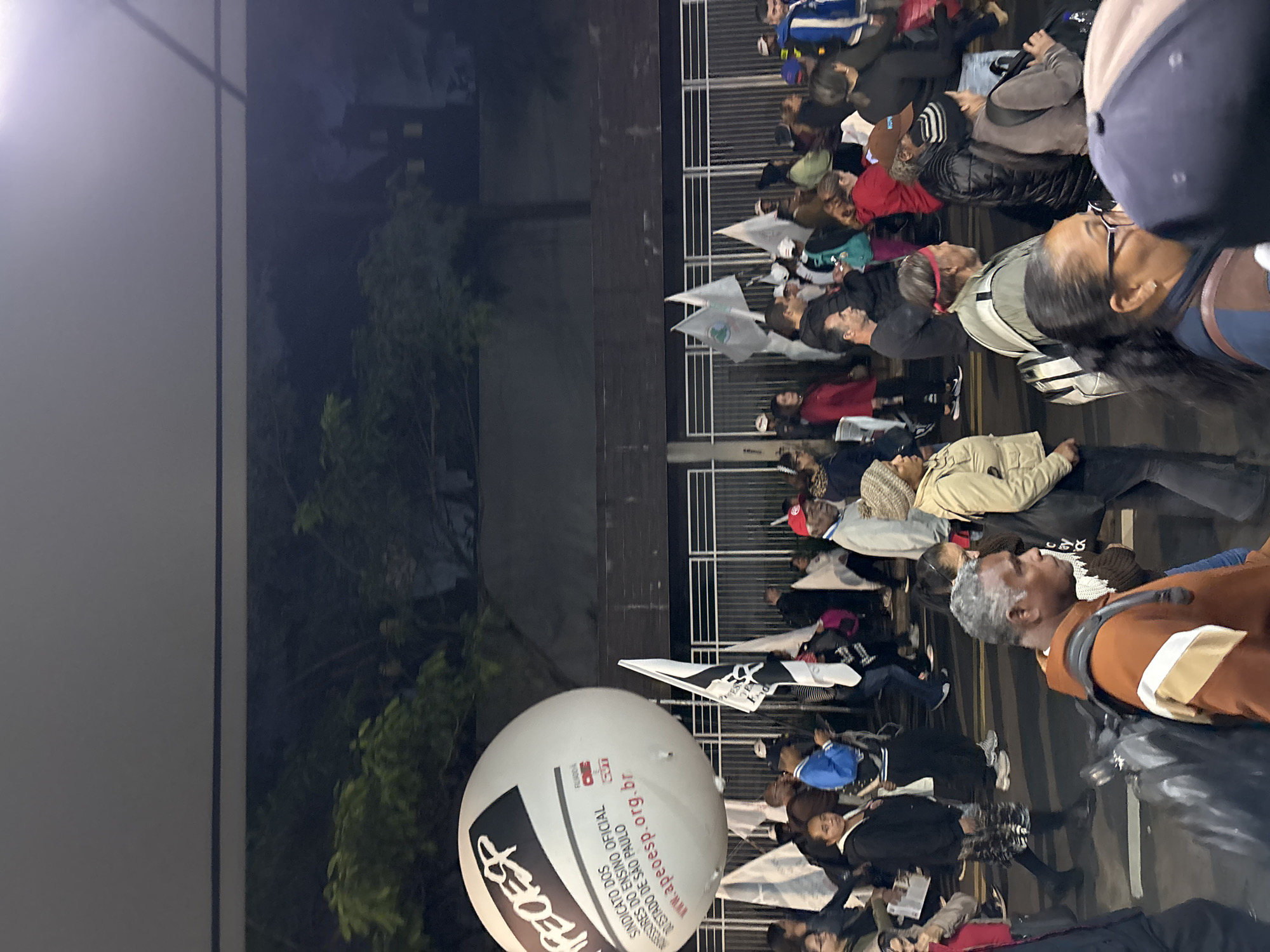 A nighttime gathering of people is underway, positioned near a dark building and adjacent greenery. The assembly appears to be a protest, indicated by the presence of flags and a large, white, spherical balloon emblazoned with "APEOESP," a union abbreviation, and web address "www.apeoesp.org.br". Most individuals are clad in casual attire, and there is an element of movement within the scene. The event appears to be in São Paulo, Brazil, judging from the union's association with the state.