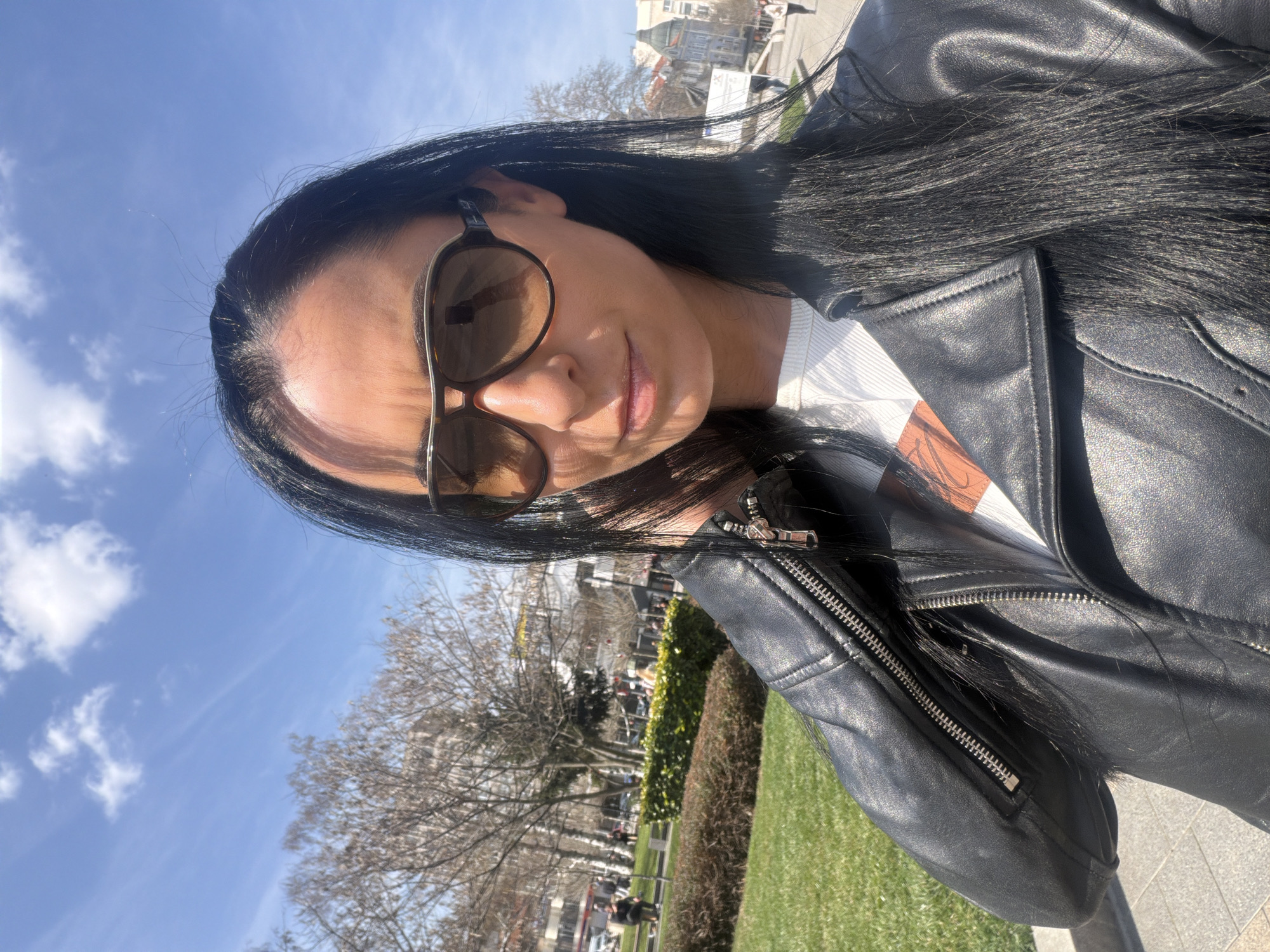 A female individual is depicted in a close-up selfie, occupying the majority of the foreground. She has long, dark hair, is wearing brown aviator-style sunglasses, a black leather motorcycle-style jacket with visible zippers, and a white ribbed shirt. Her right hand is positioned near her neck or shoulder.
The background reveals an outdoor urban park setting under a clear blue sky with scattered white clouds. To the left, a park area features green grass, bare deciduous trees, and paved walkways. Several indistinct individuals are visible walking or sitting in the distance, alongside parked cars on a street beyond the park greenery. To the right, a paved pedestrian area leads towards a street. Further back, a distinctive light-colored building with a domed roof and intricate architectural details is visible. This building is identifiable as the Regional Historical Museum, situated in Pleven, Bulgaria. The lighting indicates a sunny daytime environment.