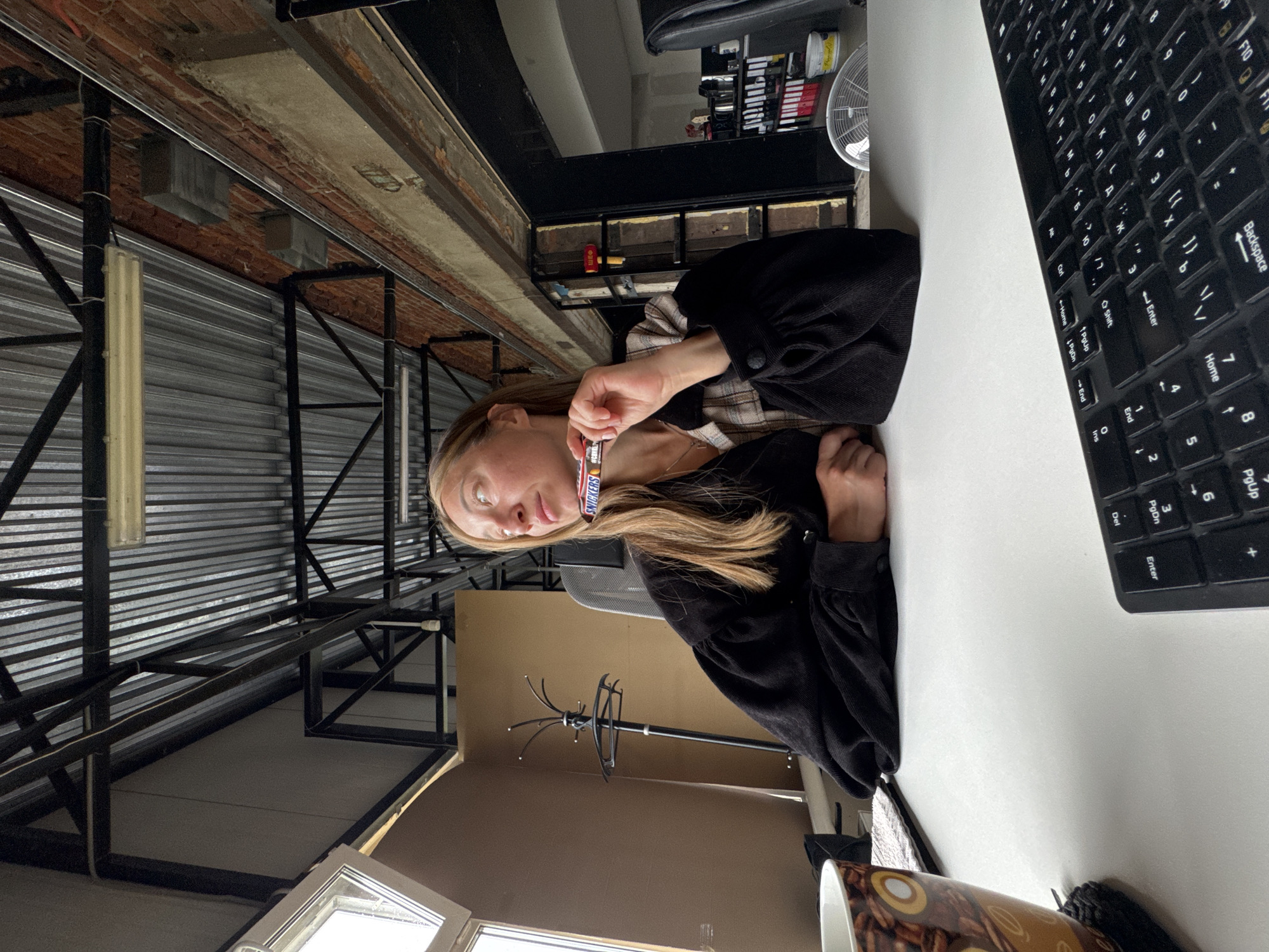 A medium shot shows a woman sitting at a white desk in what appears to be an industrial-style office space. The ceiling is corrugated metal with exposed beams and fluorescent lighting. The walls are a mix of exposed brick and painted surfaces. The woman has blonde hair and is wearing a black corduroy top with puffed sleeves and a plaid shirt underneath. She is holding a Snickers candy bar up to her mouth, looking to the left. On the desk in front of her is a coffee mug with a coffee bean pattern and a black keyboard. Behind her, a coat rack stands against a tan wall, and a window with multiple panes is visible to the left. The overall impression is a person taking a break at work in a loft-like environment. There is no information in the image to place this scene in Chaikovsky, Russia.