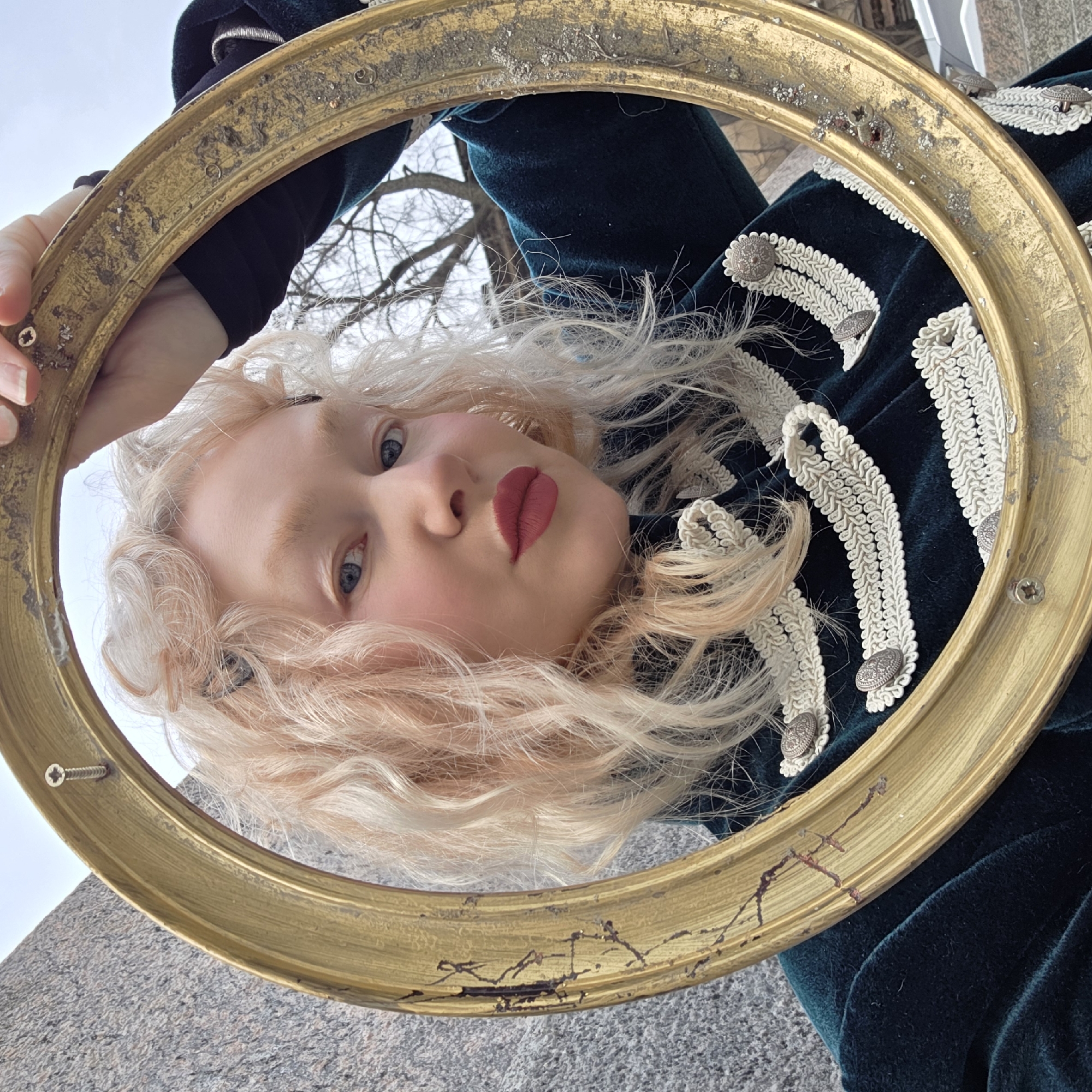 A fair-skinned person with blonde, wavy hair is holding a weathered, circular gold frame, their face centered within it. They are wearing a dark green velvet jacket adorned with white decorative trim and silver buttons. The background includes a grey stone surface and glimpses of trees and a light sky. The location is Saint Petersburg, Russia.