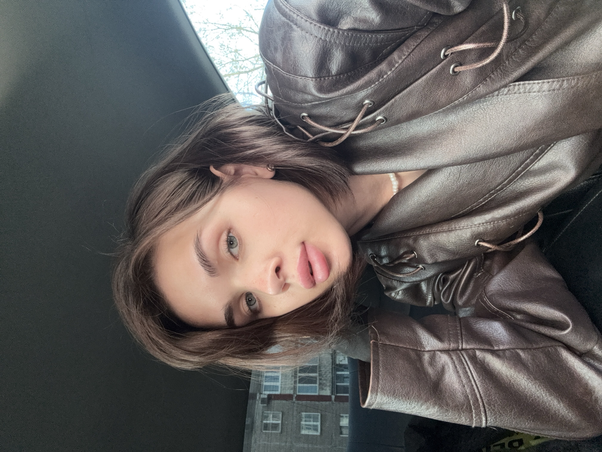 A young woman with fair skin and short, light brown hair is photographed from a low angle, appearing to be in the driver's seat of a car. She is wearing a brown leather jacket and a pearl necklace. Her mouth is slightly open, with her tongue visible. The background includes a view of a grey, multi-story building with multiple windows, suggesting an urban setting, specifically Perm, Russia. The exterior is visible through the car window. The interior of the car is dark, with a section of the dashboard visible.