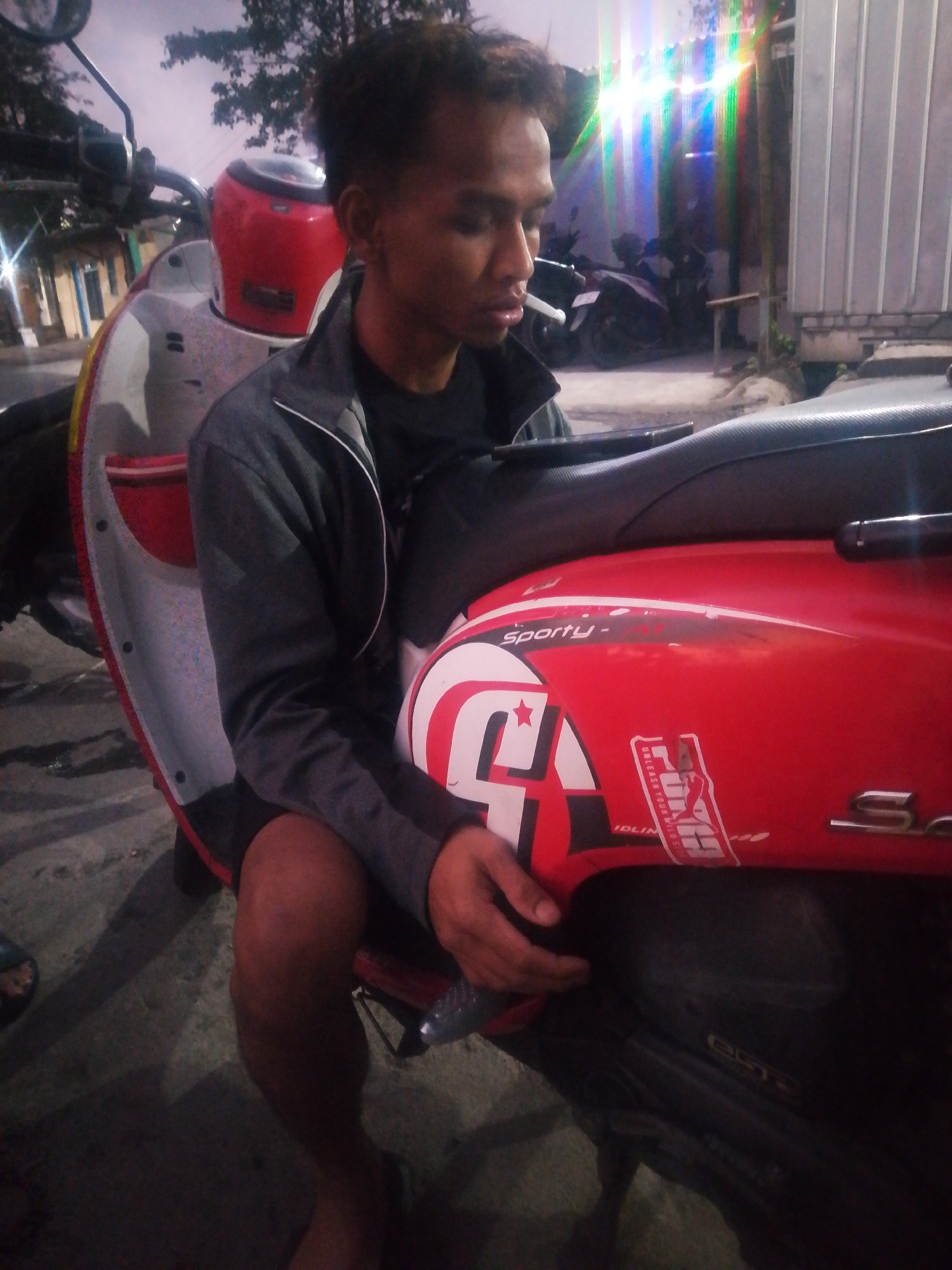 A young man is sitting on a red scooter in Tambak Sumur, Indonesia. He is wearing a dark grey jacket with white trim over a black t-shirt and dark shorts. He has short hair and is looking down at his phone. The scooter has a white and red design on its side, with the word "sporty" visible. The background shows a street with a few other parked scooters and a building. The lighting suggests it is evening or nighttime.