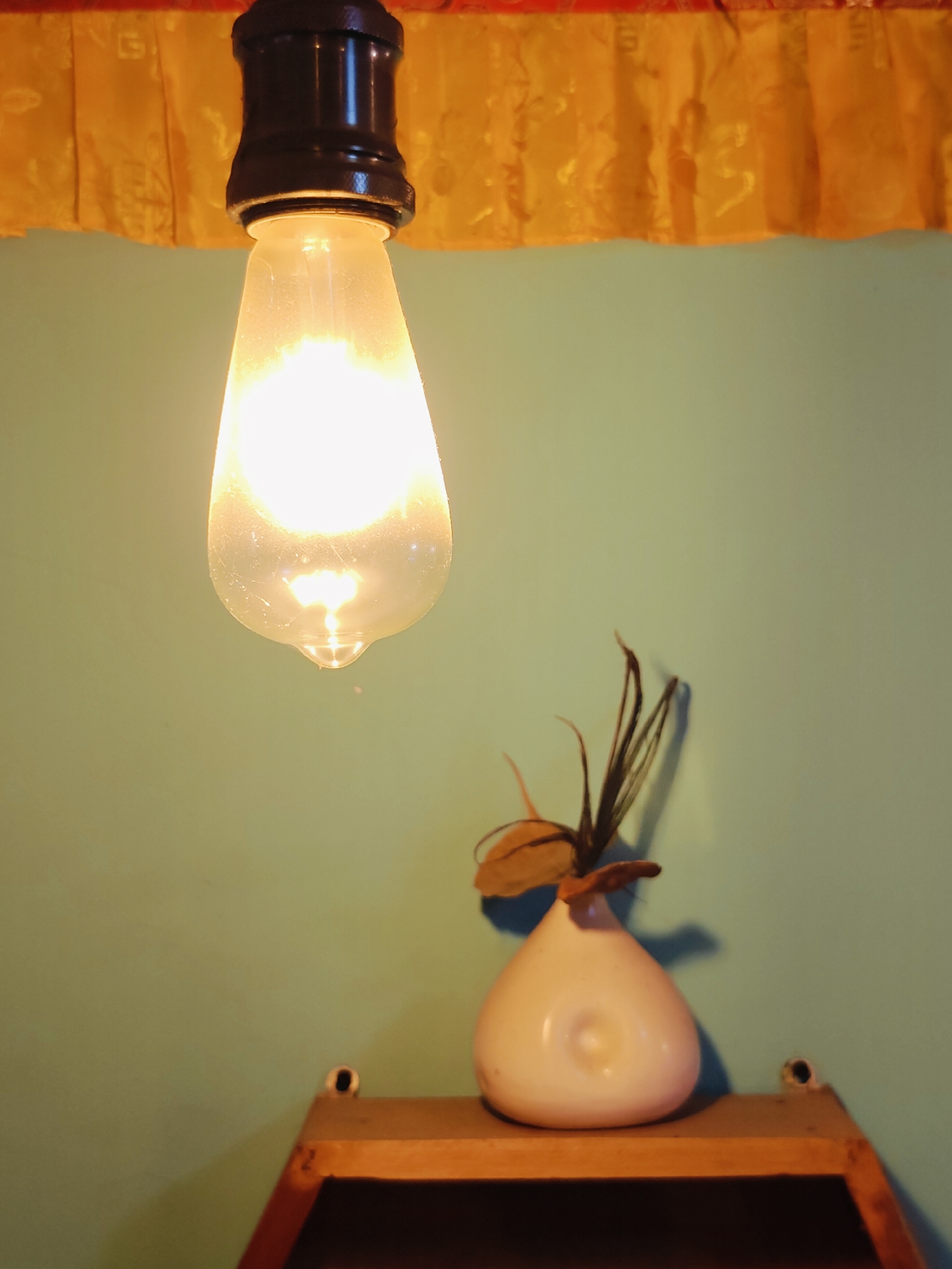 A close-up, low-angle shot captures a lit Edison-style lightbulb hanging from a dark socket against a pale blue wall. A yellow valance with a patterned texture is visible at the top of the frame. To the right, a small, white, pear-shaped vase with dried plant material sits on a wooden shelf. The shelf itself is angled and appears to be part of a larger structure, with two dark circular holes visible on its surface. The overall scene suggests an interior setting. This photograph was taken in Lalitpur, Nepal.