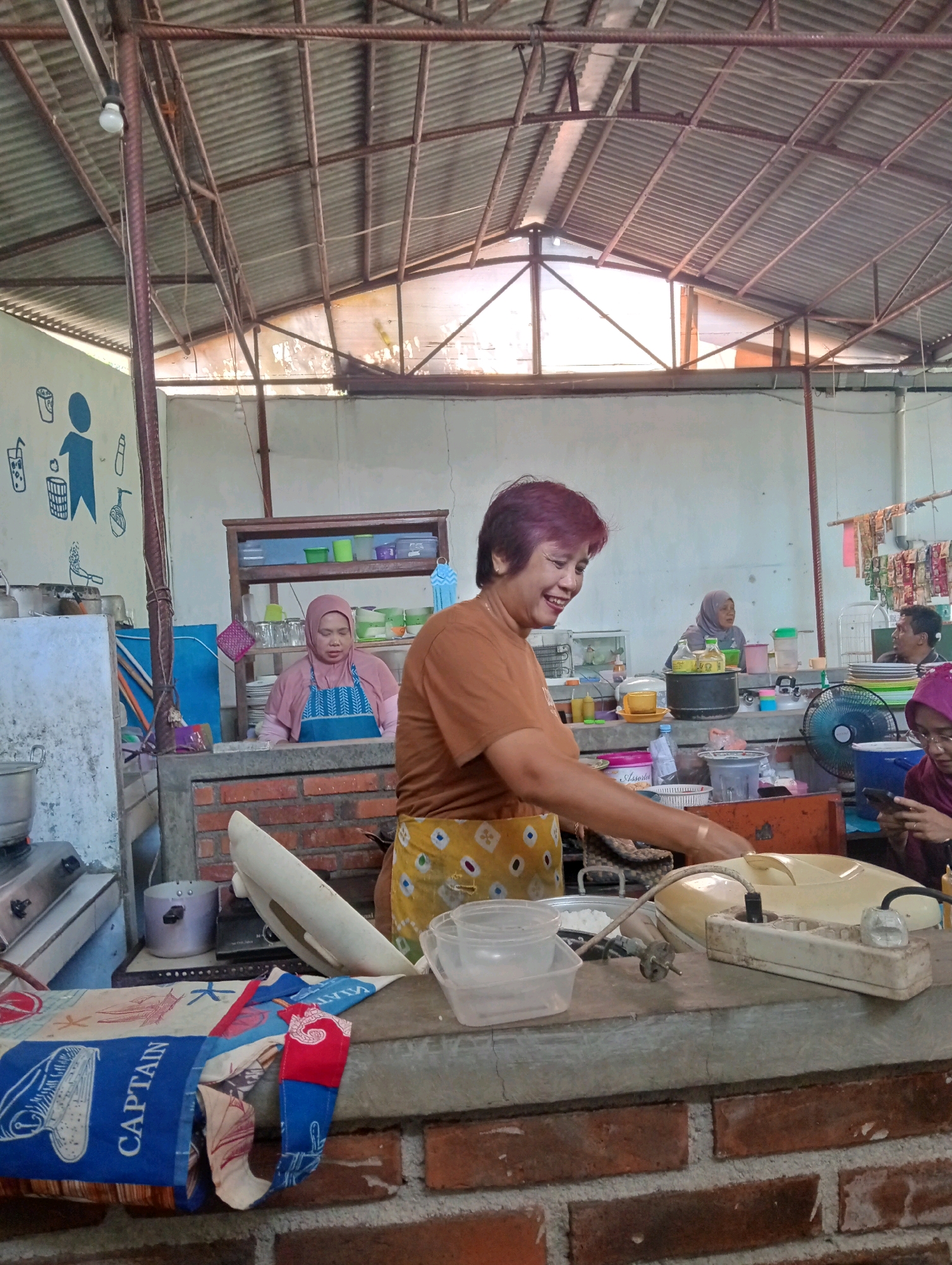 In Jampirejo, Indonesia, a woman with short, reddish-purple hair and a bright orange t-shirt is smiling and looking to her left while working at a food stall. She wears a yellow apron with a diamond pattern. The stall appears to be an outdoor or semi-outdoor establishment with a corrugated metal roof supported by metal beams. In the background, another woman wearing a pink hijab and apron is seen behind a counter, and further back, a man and another woman are visible. The stall is stocked with various food and kitchen items, including containers, plates, bottles, and cooking utensils. A fan is visible on the counter. The foreground features a brick counter and a colorful fabric with nautical designs.