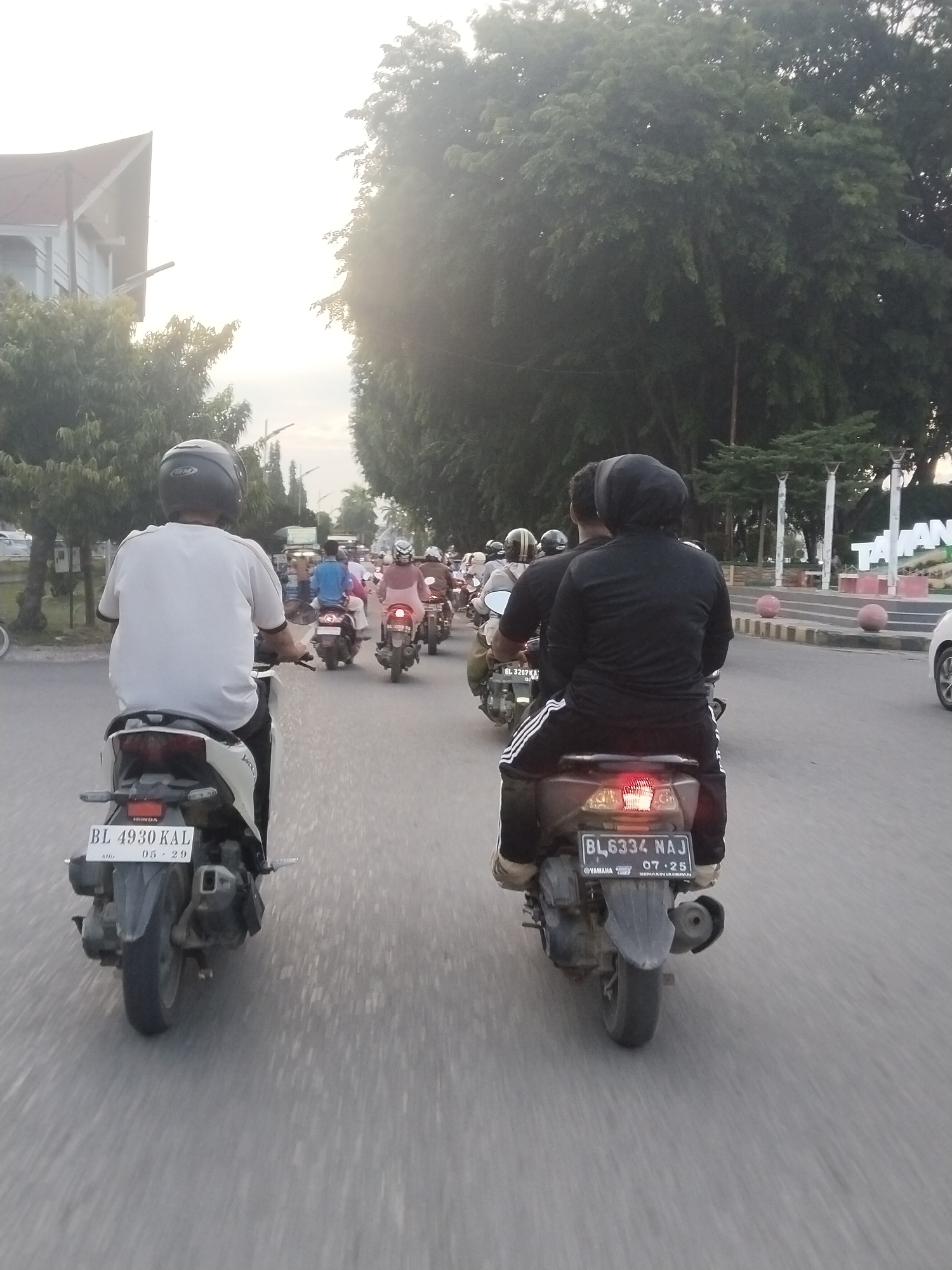 The image captures a group of people riding motorcycles on a paved road in Lhokseumawe, Indonesia. The perspective is from behind one of the riders, looking forward along the road. In the foreground, a person wearing a white t-shirt and a black helmet rides a light-colored motorcycle with a license plate that reads "BL 4930 KAL 05 29". To the right, another motorcycle carries two individuals; the passenger, in the foreground, wears a black headscarf and a black outfit with white stripes on the pants, and the motorcycle's license plate is "BL 6334 NAJ 07 25". Several other motorcycles and riders are visible further down the road, some in motion, forming a convoy. The environment includes trees lining the road, a building with a pitched roof on the left, and a distant structure with the word "TAH" visible, possibly part of a sign. The sky is overcast with bright light suggesting late afternoon or early morning. The road surface shows motion blur, indicating the motorcycles are moving.