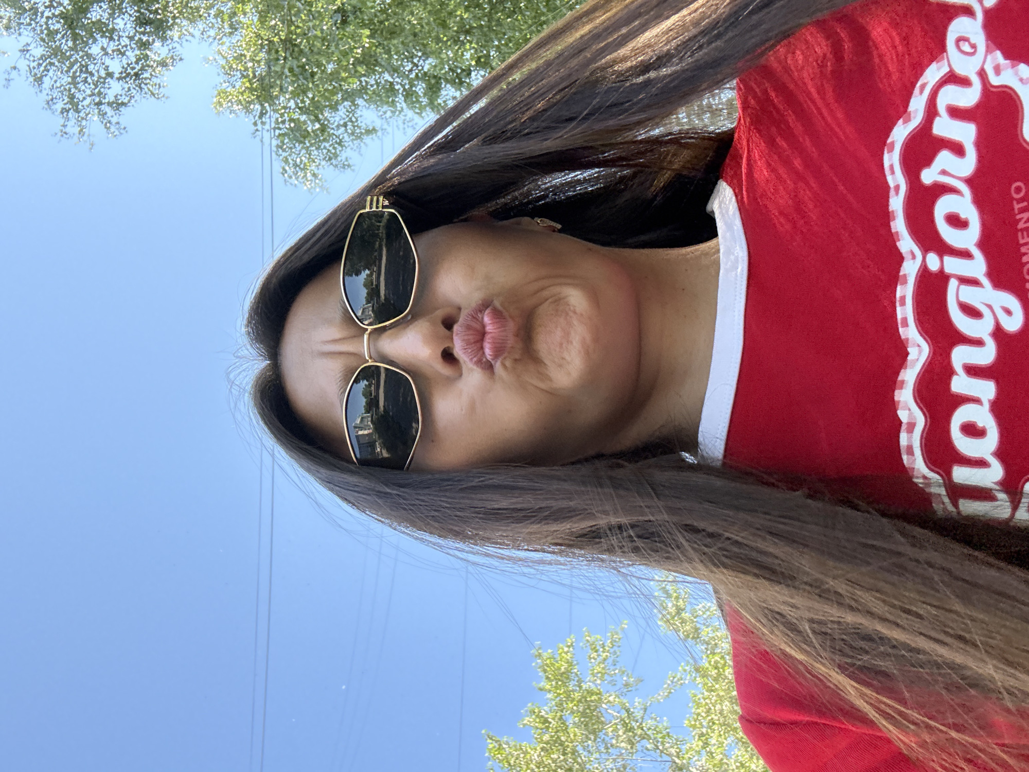 A low-angle, close-up, outdoor selfie features a young woman with long, dark brown hair, wearing sunglasses and a red t-shirt. Her lips are pursed in a kissing gesture. The sunglasses reflect a street scene with buildings and trees. The background consists of a clear blue sky with some power lines and green trees on either side. The t-shirt has a white collar and a graphic design that includes the word "Buongiorno" in white lettering. There are no discernible elements that suggest the location is Chaikovsky, Russia.