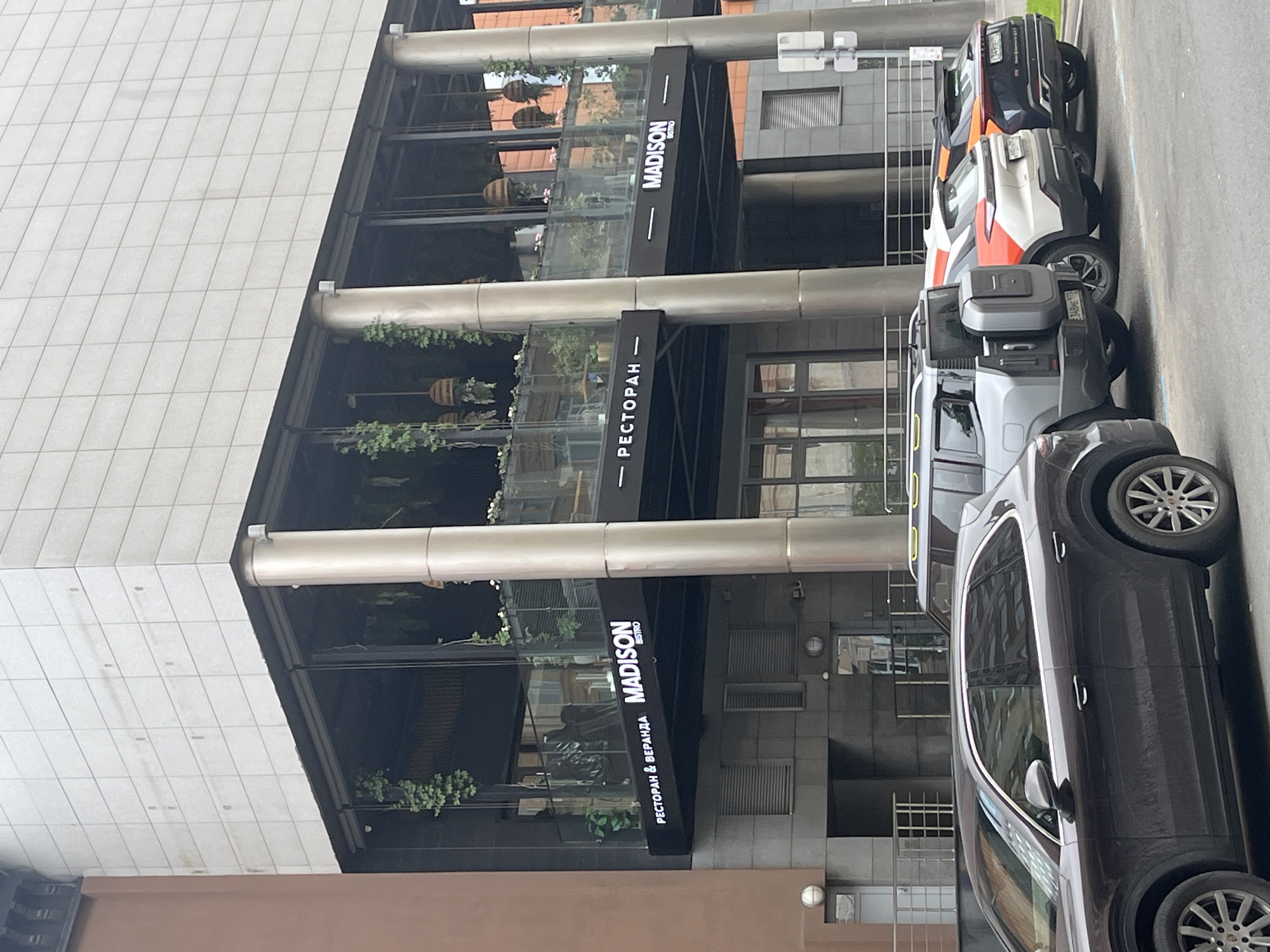 A dark brown SUV is parked in the foreground, facing right. Behind it, a silver SUV is parked, also facing right. To the right of the silver SUV are two more vehicles, one white with orange accents and the other dark blue, both facing right. The vehicles are parked in front of a modern building with a glass-enclosed second-floor terrace. The building's facade is made of light-colored tiles, and it has several large, circular pillars supporting the second floor. A sign on the building reads "MADISON BISTRO" and "РЕСТОРАН & ВЕРАНДА". Greenery hangs from the terrace railing and from the ceiling of the terrace. The environment appears to be an urban setting, with a cloudy sky overhead.