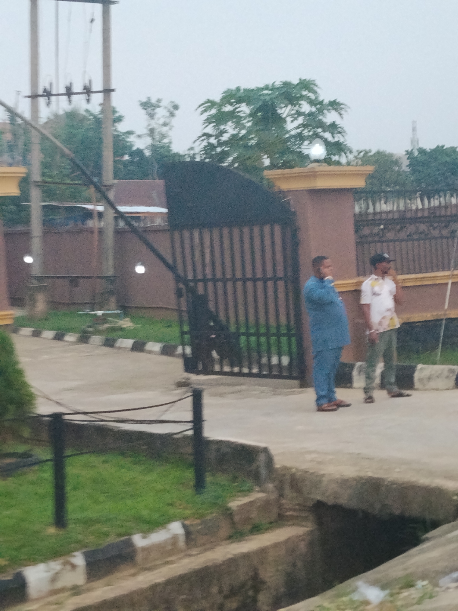 The image depicts two men standing near a gate in Ura Nta Umuarandu, Nigeria. The man on the left is wearing a blue outfit, while the man on the right is wearing a white shirt, green pants and a baseball cap. Behind them is a brown fence with a black metal gate. The setting appears to be a residential or commercial area, with trees visible in the background. In the foreground there is a grassy area with concrete borders and a drainage ditch. Utility poles are also visible in the background. The sky is overcast, suggesting it may be early morning or late afternoon.