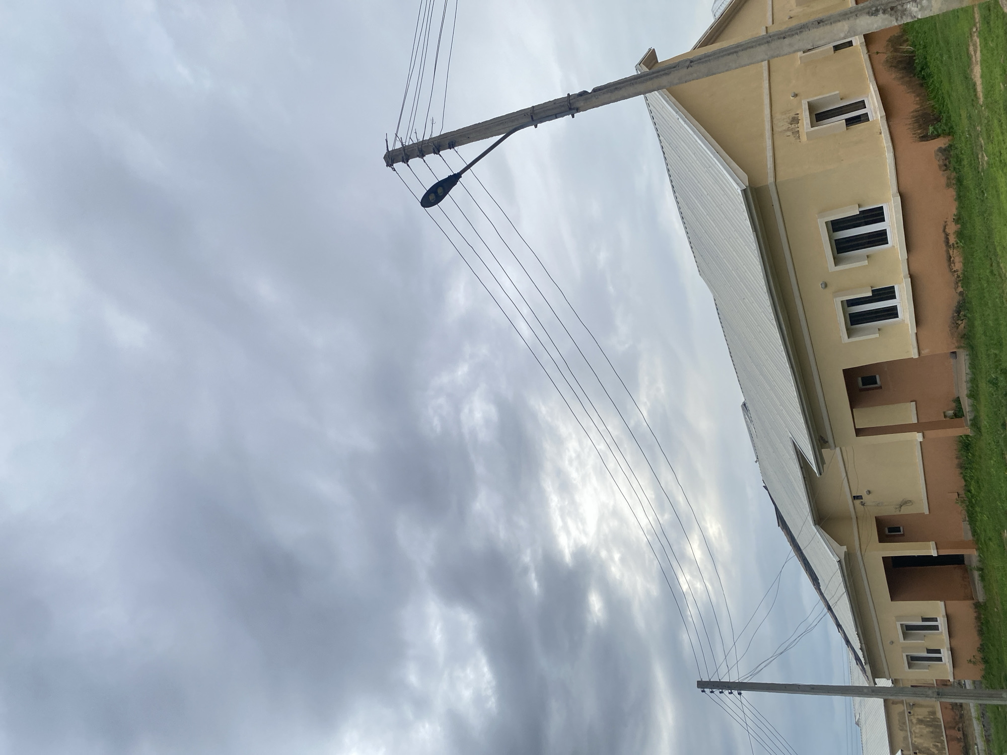 A low-angle, vertical, full shot captures a street scene under a heavy, overcast sky, with light filtering through broken clouds. In the foreground, a concrete utility pole with a street lamp attached stands on the right, supporting multiple electrical wires that crisscross the frame. To the left, another, thinner utility pole with insulators is visible. The middle ground features a row of tan and light orange single-story houses with white roofs. The houses have dark-framed windows and appear to be under construction or newly built, with some visible staining on the lower exterior walls. A patch of green grass is in front of the houses. The sky dominates the upper portion of the image with its dense, gray cloud cover, hinting at approaching rain.