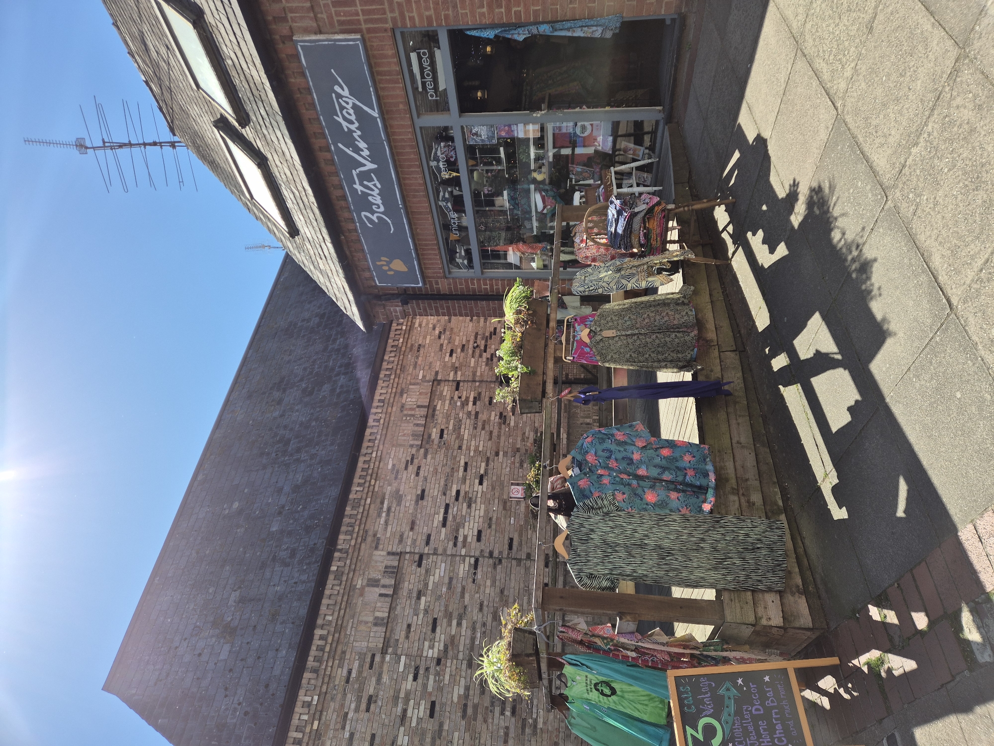 This exterior shot captures a bright, sunny daytime scene in front of "3 Cats Vintage," a shop likely situated in West Suffolk, United Kingdom. The building features a red brick facade with a dark tiled roof and visible skylights. A prominent antenna structure rises against the clear blue sky above the building.
The shop's main sign, a dark rectangle with white cursive lettering, reads "3 Cats Vintage" and includes a paw print logo. The large display window shows interior merchandise and bears decals stating "preloved," "retro," and "unique."
In front of the shop, an outdoor display showcases various items. Garments with colorful patterns, including dresses and tops, hang on wooden racks. Planters with greenery are integrated into the display, adding a natural touch against the brick wall. A small A-frame chalkboard sign reiterates "3 Cats Vintage" and lists its offerings: "Clothes," "Jewellery," "Home Decor," "Charm Bar," and "and much more!"
The ground is a mix of paved flagstones and some brick sections. Strong, angular shadows are cast across the walkway, indicating direct sunlight from a relatively high angle. The overall impression is that of a charming and inviting local business, bathed in bright sunshine. No people are visible in the image.
