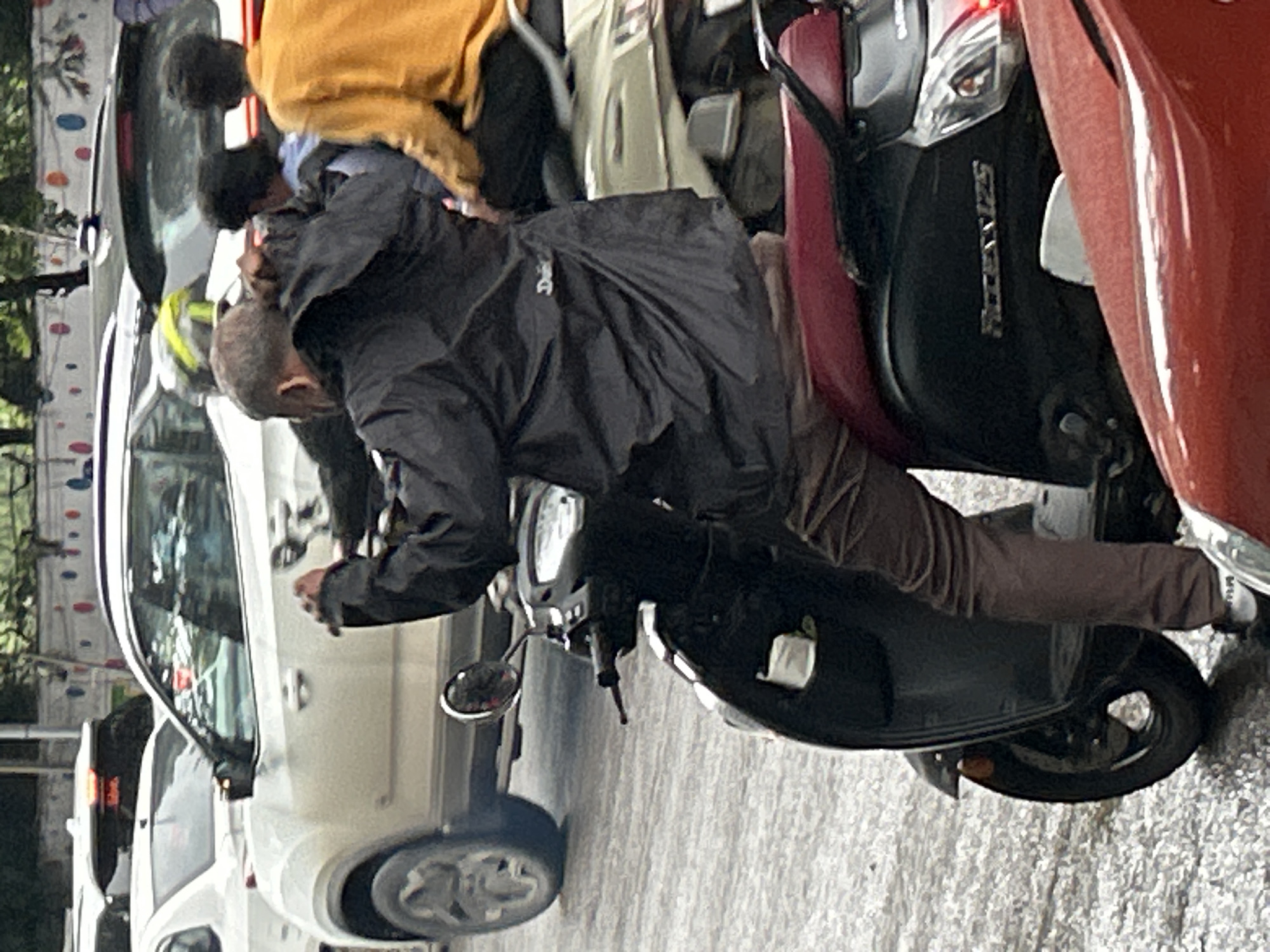 This is a medium shot of a traffic scene in Pune, India, on a wet or damp day. In the foreground, a person wearing a black hooded jacket and brown pants is sitting on a black scooter. The scooter has red accents on the seat and a silver "Access 125" emblem on the rear panel. The person is holding onto the handlebars, and their jacket appears to be slightly damp. Behind the scooter, another person wearing a bright yellow long-sleeved shirt is seated on a lighter colored scooter, and further behind them, a third person is partially visible. In the background, several white cars are present, including what appears to be an SUV. The road surface is wet and reflective. The scene suggests a busy urban environment with typical traffic conditions in Pune.