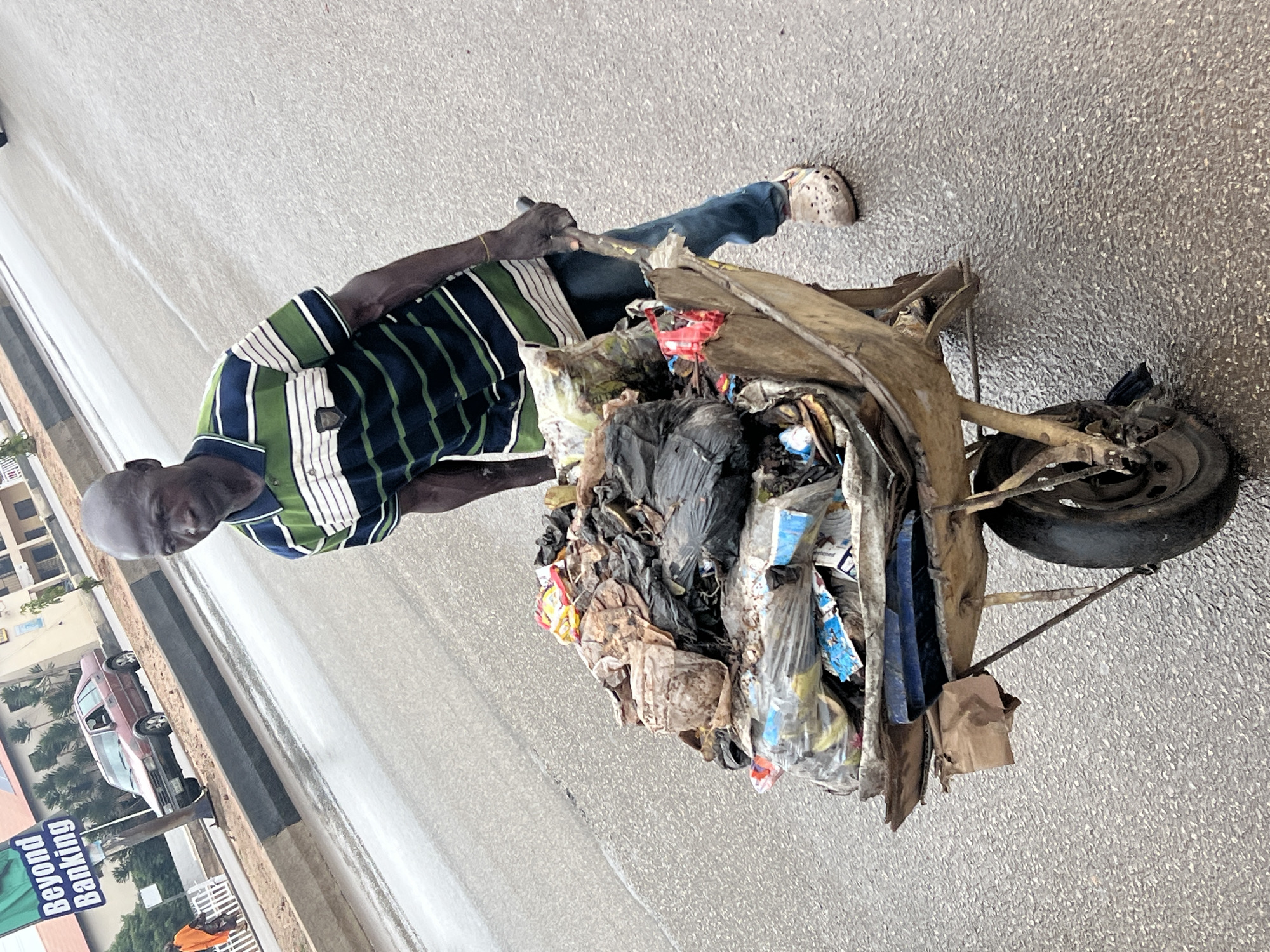 A man is pulling a wooden, metal-wheeled cart overflowing with trash on a paved street in Gura Top, Nigeria. The man wears a horizontally striped green and blue shirt, jeans, and beige clogs. The cart contains black plastic bags, clear plastic bags with colorful items, and other refuse. The pavement is gray. A building, car, and a large advertising sign are visible in the background.