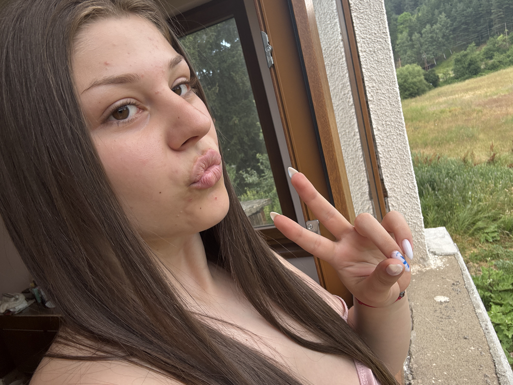 A young woman with long brown hair is shown in a close-up, selfie-style shot, making a kissy face and holding up a peace sign. She is standing on a balcony overlooking a grassy field and a wooded area. The woman is wearing a pink top and has a red string bracelet on her wrist. Her nails are painted with white and blue designs. The background shows a rural landscape with trees and hills. The lighting suggests it is daytime.