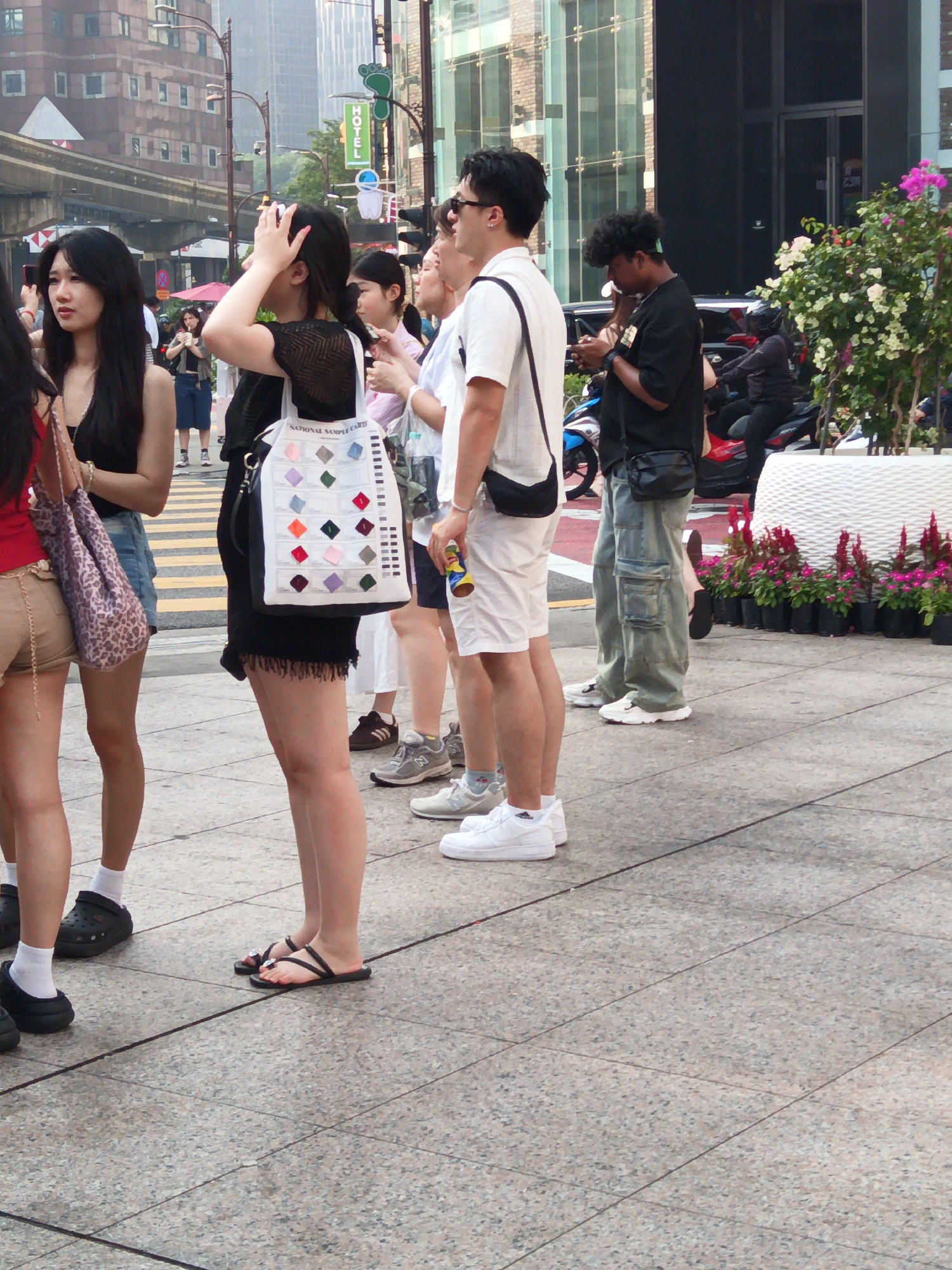 A medium shot captures a group of individuals standing on a paved sidewalk in Kuala Lumpur, Malaysia. The scene appears to be a commercial or pedestrian area with a blurred urban background that includes buildings, a monorail track, and streetlights. In the foreground, several people are standing and walking. On the left, a woman with long dark hair is wearing a black top and a denim skirt, carrying a patterned handbag. To her right, a woman in a black top and a black skirt holds a white tote bag with colorful squares. Behind them, a woman with dark hair is adjusting her hair, and another woman in a white shirt is visible. Further back, a man in a black t-shirt and denim pants is looking at his phone, and another man in a white shirt and shorts is holding a drink can. The ground is a light-colored tiled pavement. In the background, there are planters with pink and red flowers.