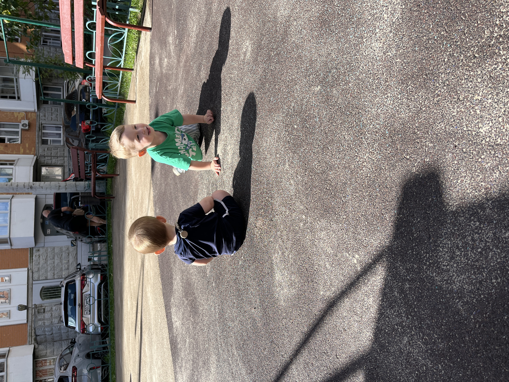 Two young children are on a paved outdoor area, likely a playground or courtyard, in Balashikha, Russia. The child on the left, facing away from the camera, is sitting down and wearing a dark blue shirt and shorts. They have a wooden beaded necklace. The child on the right is on their hands and knees, wearing a green t-shirt and striped shorts. They are looking to their left. In the background, a building with multiple windows and balconies is visible, along with parked cars, a metal fence, and two red benches. The ground is dark asphalt or a similar material with visible specks. Shadows cast by the children and unseen objects are prominent on the ground.