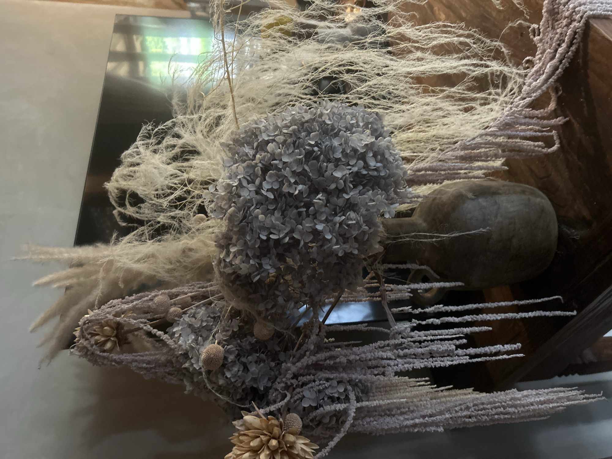 This close-up image presents an intricate decorative arrangement predominantly featuring various dried and preserved botanical elements. At the core are several large, spherical clusters of dried hydrangea blossoms, characterized by their muted grey-blue hue and delicate, papery petals. These are surrounded and interwoven with abundant quantities of light-colored, wispy dried grasses or reeds, which create a soft, ethereal texture and extend broadly across the frame.
Numerous stringy, elongated plant elements, possibly dried amaranth or similar, with a granular or fuzzy texture and in shades of light brown and dusty pink, are also prominent. Smaller, spiky seed pods and dried flower heads further diversify the botanical display. A dark, gourd-like object or ceramic vessel is partially visible on the right side, integrated into the rustic composition.
The background includes reflections on a dark, glossy surface, likely a mirror or polished table, which shows blurred natural light from an unseen window, indicating an indoor setting during daytime. The arrangement evokes a sense of natural beauty and tranquility, emphasizing diverse textures and muted color tones. No specific activity is taking place, and no text is visible.