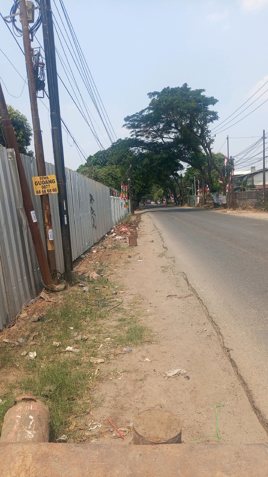The image shows a street scene in Kadu, Indonesia. On the left side of the image, a tall metal fence runs along the side of the road, bordering what appears to be a construction site or undeveloped land. In front of the fence are utility poles with a tangle of wires. A yellow sign is attached to one of the poles, which reads "SEWA GUDANG" (Warehouse Rental) and provides a phone number. The ground next to the fence is covered with dry grass, dirt, and scattered debris, including plastic bottles and paper.
The road itself is asphalt and appears to be two lanes wide, curving slightly to the right. Along the side of the road, there is a dirt and gravel shoulder with several circular concrete or metal structures partially embedded in the ground, possibly manhole covers or stubs. There are also some small concrete posts lining the shoulder.
In the distance, trees with dense green foliage arch over the road, creating a shaded canopy. Small flags, possibly Indonesian flags given the red and white colors, are visible hanging from poles along the roadside on the right. Buildings and more trees are visible further down the road on the right. The sky is clear and blue. Two large rusty cylindrical objects are in the foreground on the lower left, one of which has some markings on it. Another circular object is in the foreground on the lower right.