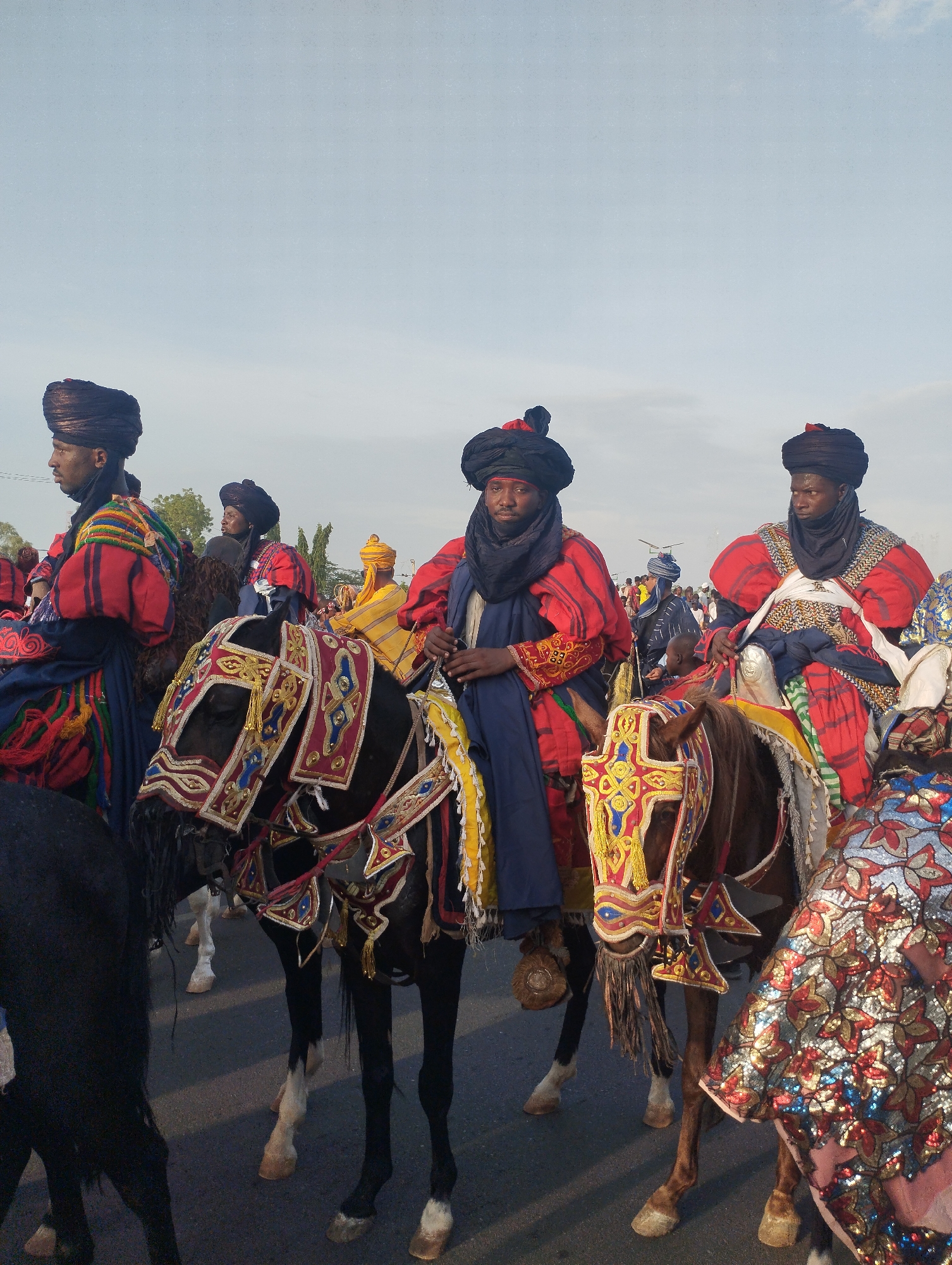 In the city of Gombe, Nigeria, a procession of men on horseback is captured. The riders are adorned in vibrant red and blue traditional attire, complete with intricately wrapped turbans. The horses are equally embellished with colorful, ornate tack. The sky is a pale blue, suggesting either early morning or late afternoon light. The scene is likely a cultural event or festival.