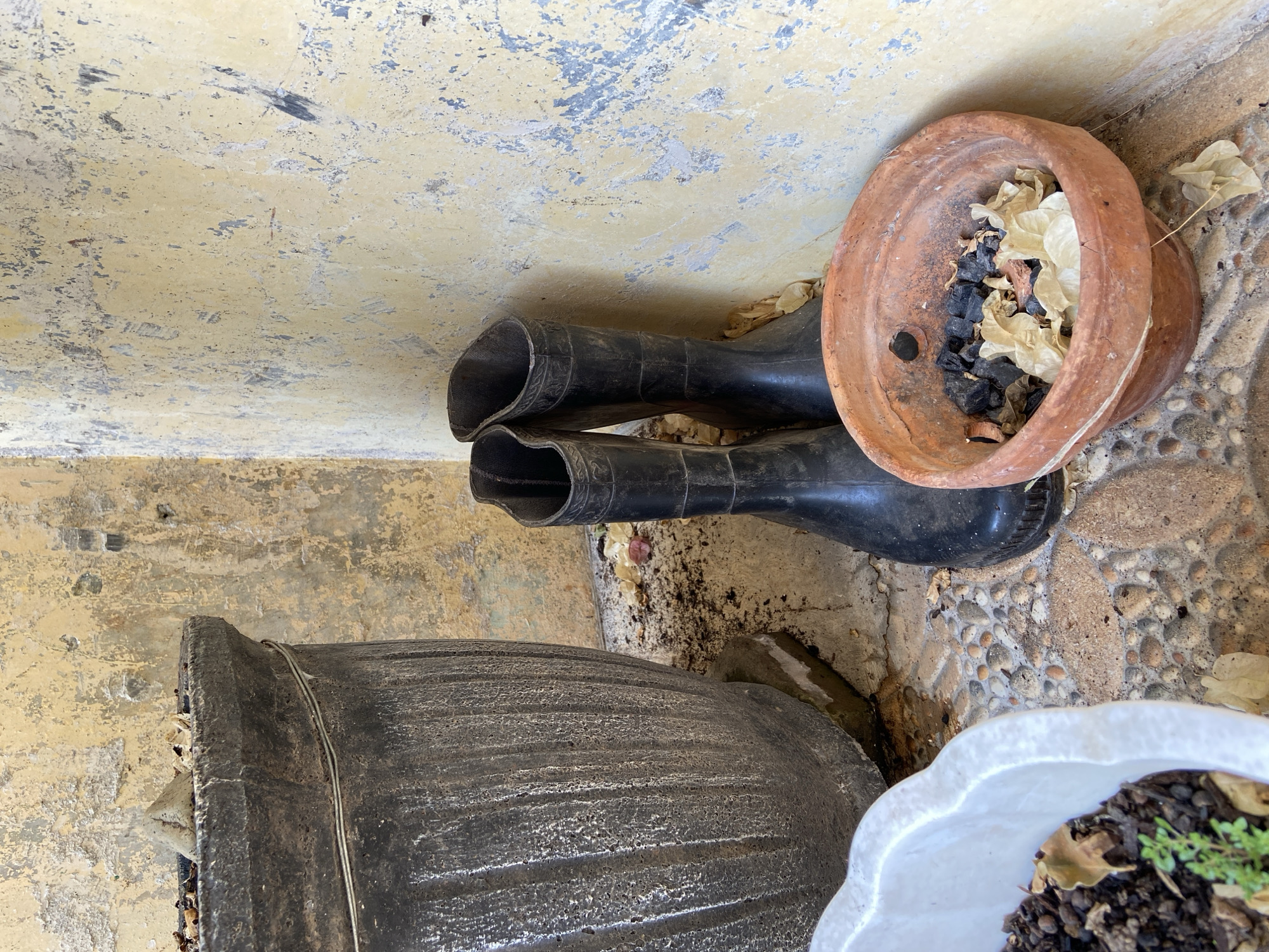 Two black rubber boots stand upright in a corner formed by a weathered, yellow-painted wall and a large, dark gray planter. The boots are positioned close together, with the wall behind them. In the foreground, a terracotta pot filled with charcoal and dried leaves sits to the right of the boots. To the left, a portion of a white planter with soil and greenery is visible. The ground is a mosaic of small, round stones. The overall scene appears to be an outdoor storage or utility area. The location is Margorejo, Indonesia.