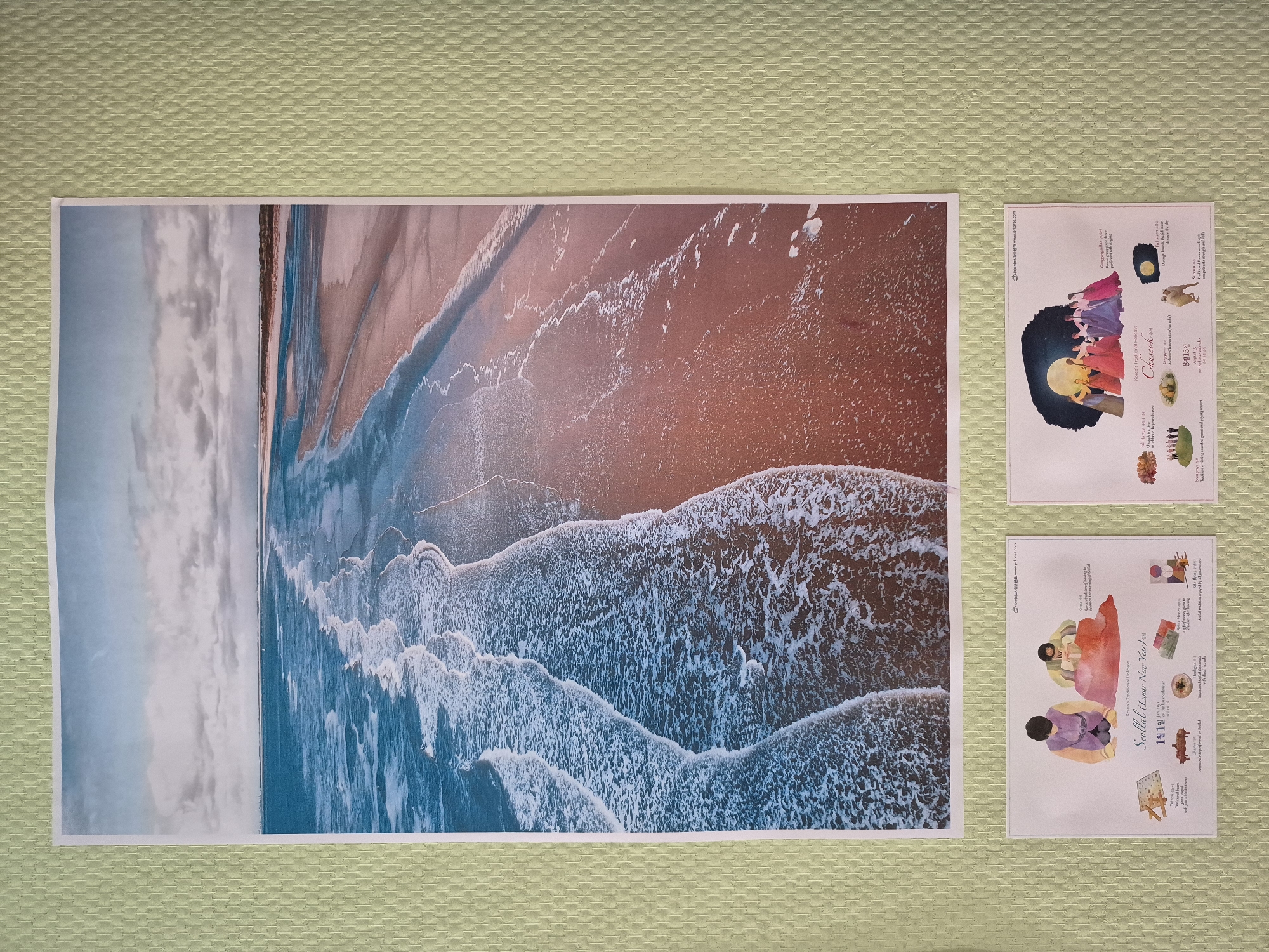 The image displays a triptych of Korean cultural information posters mounted on a textured light green wall. The central and largest poster features an aerial view of a sandy beach with waves crashing on the shore. The smaller posters below are informational graphics detailing Korean holidays. The left poster, titled "Seollal (Lunar New Year)", depicts two people in traditional Korean attire performing a ritual and illustrates related traditions such as a board game, ancestral rites, a traditional dish, and a gift box. The right poster, titled "Chuseok", shows a group of people in traditional dress gathered under a full moon and describes related traditions like a harvest celebration, ancestral rites, and a depiction of people offering food to the moon. There is no indication of the city of Moskva, Russia, in this image.