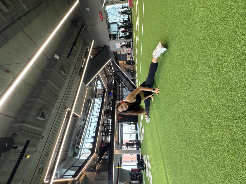 A young adult female is centered in a modern, multi-level fitness center in Bangkok, Thailand, performing a full straddle split on a green artificial turf floor. She wears a black sports bra, black leggings, white athletic shoes, a dark smartwatch on her left wrist, and red earbuds. Her hands are placed flat on the turf in front of her, and she looks directly forward.
The environment features an exposed concrete ceiling with linear LED lighting fixtures and visible ventilation ducts. Black metal railings delineate upper mezzanine levels, where multiple individuals are visible utilizing cardio equipment such as treadmills. On the lower level, a red sign displaying "BODY CAMP" and a red square marked with the number "1" are visible. A digital clock on a gray wall in the background shows "16:03:48." Other gym equipment, including weight racks and large training tires, are present in the mid-ground and background. Large windows extend along the far side of the facility, revealing exterior urban buildings.