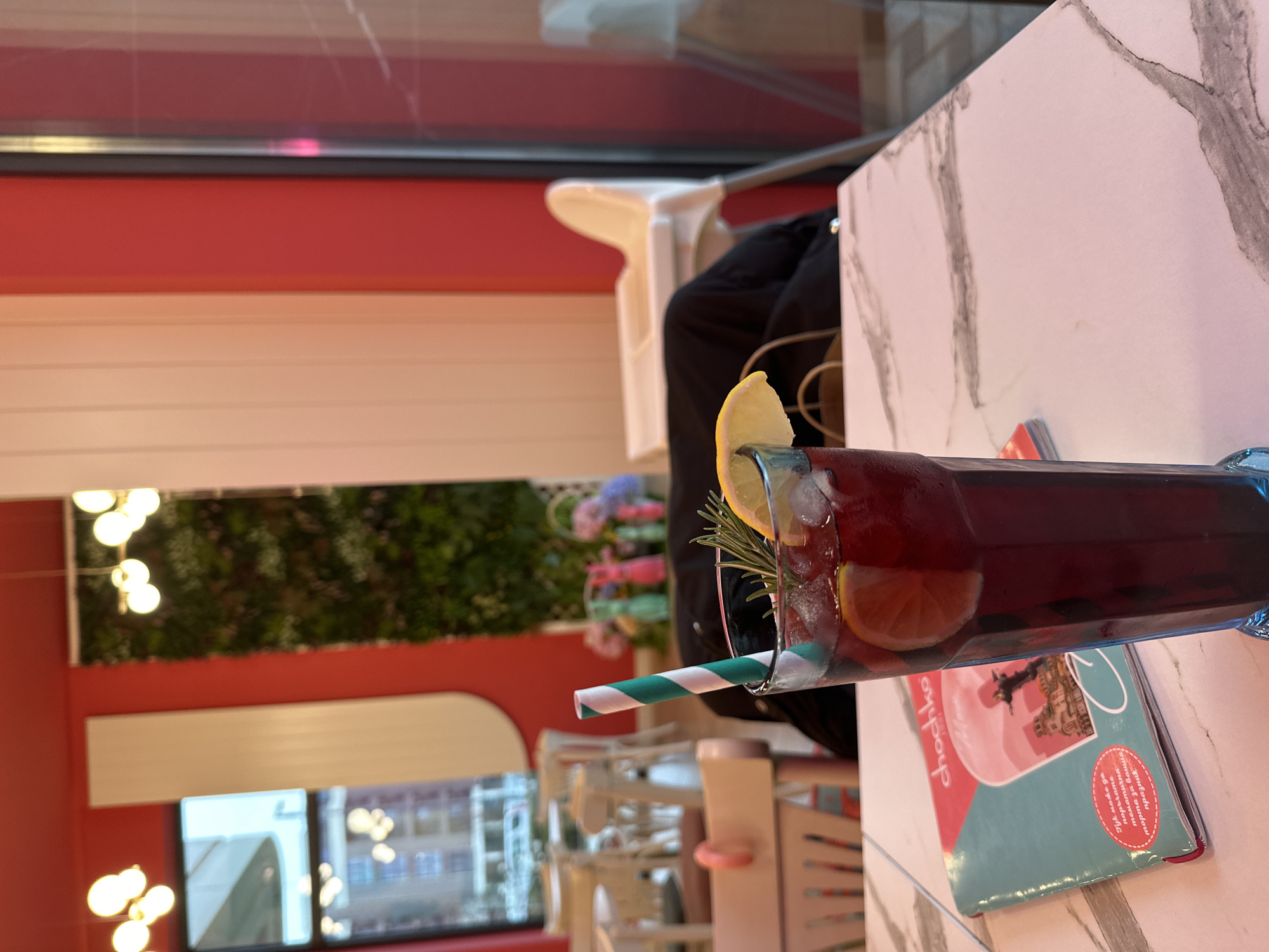 A close-up shot of a beverage served in a glass with ice, lemon slices, and a green-and-white striped straw. The glass sits on top of a book or magazine on a table.
The setting appears to be inside a cafe or restaurant, with white chairs and other tables visible in the background. A window provides a view of buildings outside, suggesting an urban location.
Given the information on the book and the cafe setting, it's highly likely that the location is in Sofia, Bulgaria.