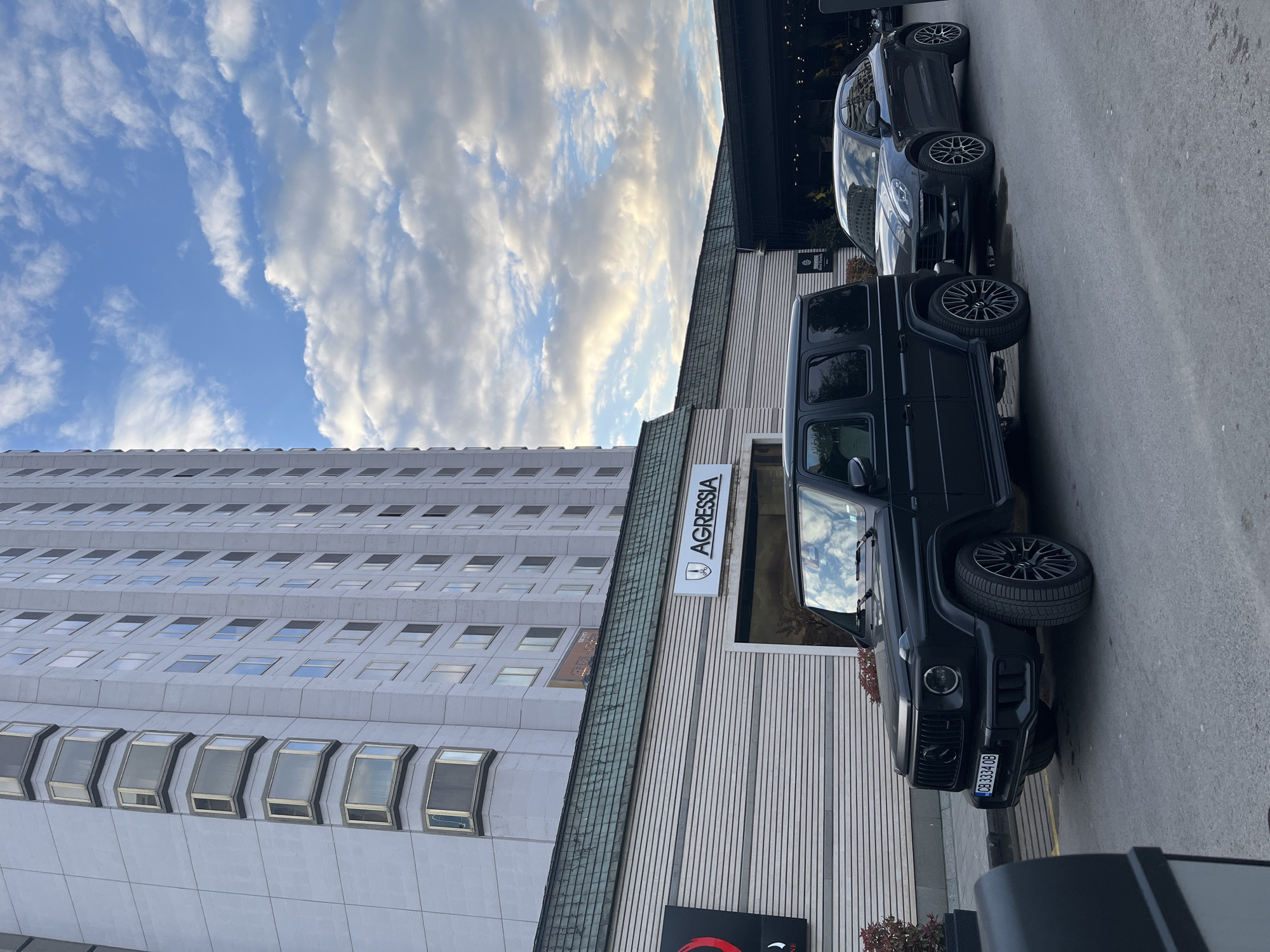 This image captures an urban street scene in Sofia, Bulgaria, on a partially cloudy day.
In the foreground, two dark-colored luxury SUVs are parked alongside a building. The vehicle closer to the viewer is a black Mercedes-Benz G-Wagon (G-Class), prominently displaying a Bulgarian license plate "CB 3334 OB". Its dark wheels and rugged design are visible, with reflections of the sky and clouds in its windows. Behind the G-Wagon, another dark SUV, possibly a Mercedes-Benz as well, is partially visible.
To the right of the parked vehicles stands a modern building with light-colored, vertically slatted siding. A prominent sign on this building reads "AGRESSIA," accompanied by a circular logo.
To the left, a tall, multi-story building of light grey color rises into the background. This building features a distinctive facade with numerous small, rectangular windows arranged in horizontal rows across its many floors. A faint "SEIKO" text can be discerned on a billboard or sign on the side of this tall building.
The sky above is bright blue with scattered white, fluffy clouds, suggesting clear weather during daytime, likely late morning or afternoon. The cars are stationary, indicating they are parked in an urban environment.