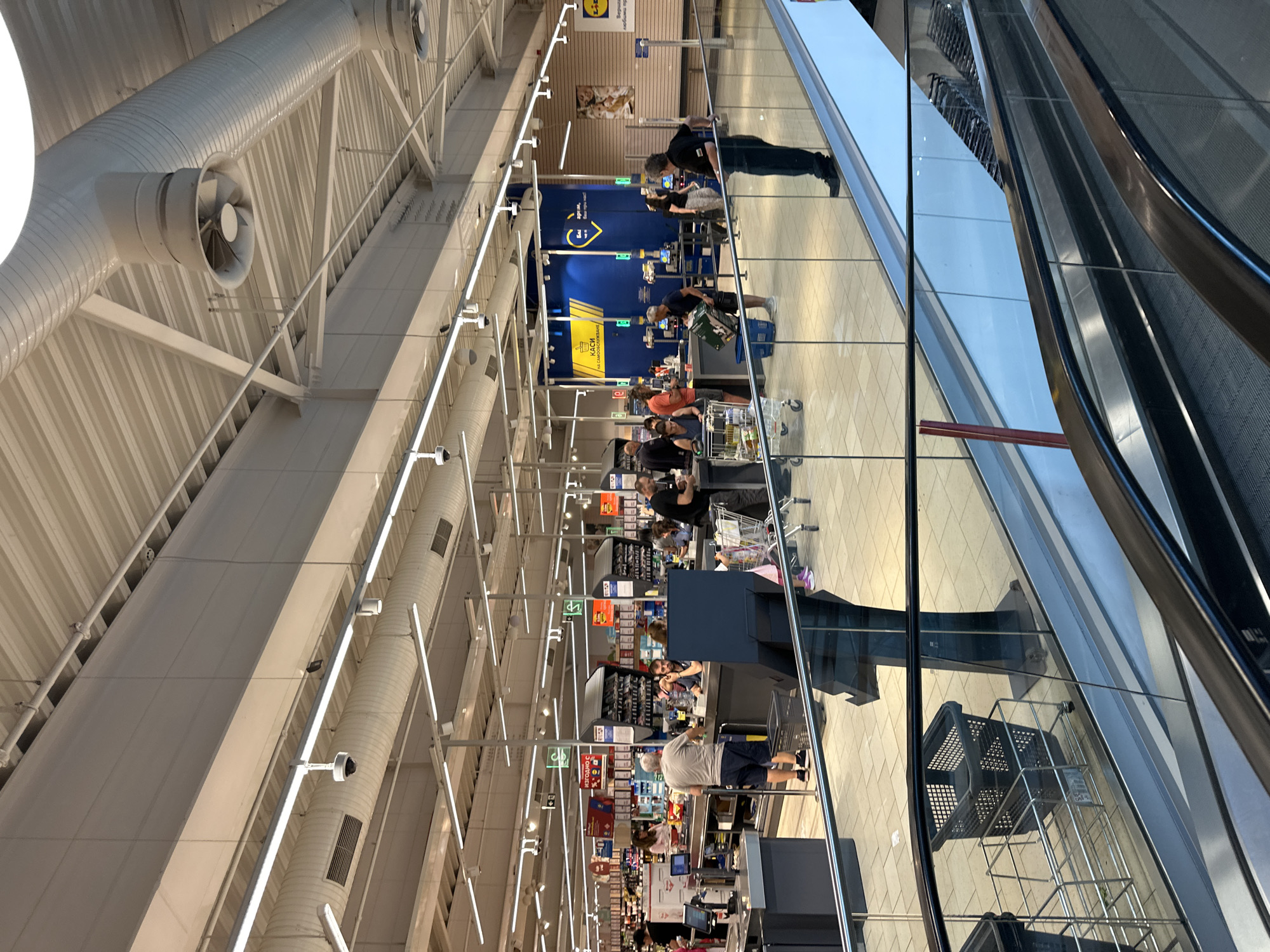 The image captures the interior of a modern supermarket, likely a Lidl given the visible branding on the right side of the image and on some signage. The scene is set within the city of Sofia, Bulgaria. In the foreground, a portion of an escalator with a glass railing is visible, suggesting a multi-level retail space or an entrance area.
The main focus of the image is the checkout area, where several customers are interacting with cashiers. There are multiple checkout lanes, some of which are occupied. People are seen with shopping carts and baskets, indicating they are in the process of purchasing items. The environment is brightly lit with fluorescent lighting, and ventilation ducts are prominently visible along the ceiling. Security cameras are also mounted on the ceiling. The flooring appears to be tiled. The overall atmosphere is that of a busy commercial space.