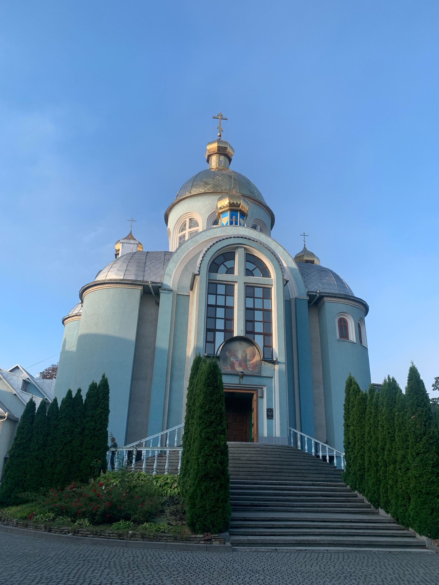 The image shows an exterior view of a light blue Orthodox church with golden domes, located in Kolomyia, Ukraine. The church features a prominent set of wide stone stairs leading up to an arched wooden entrance. White balustrades line the sides of the staircase, and a colorful religious mural is visible above the entrance. Tall, slender green cypress trees flank the stairs, and a garden with red roses and other foliage is situated to the left. The sky is a clear, bright blue, suggesting it is daytime. The foreground is paved with cobblestones.
