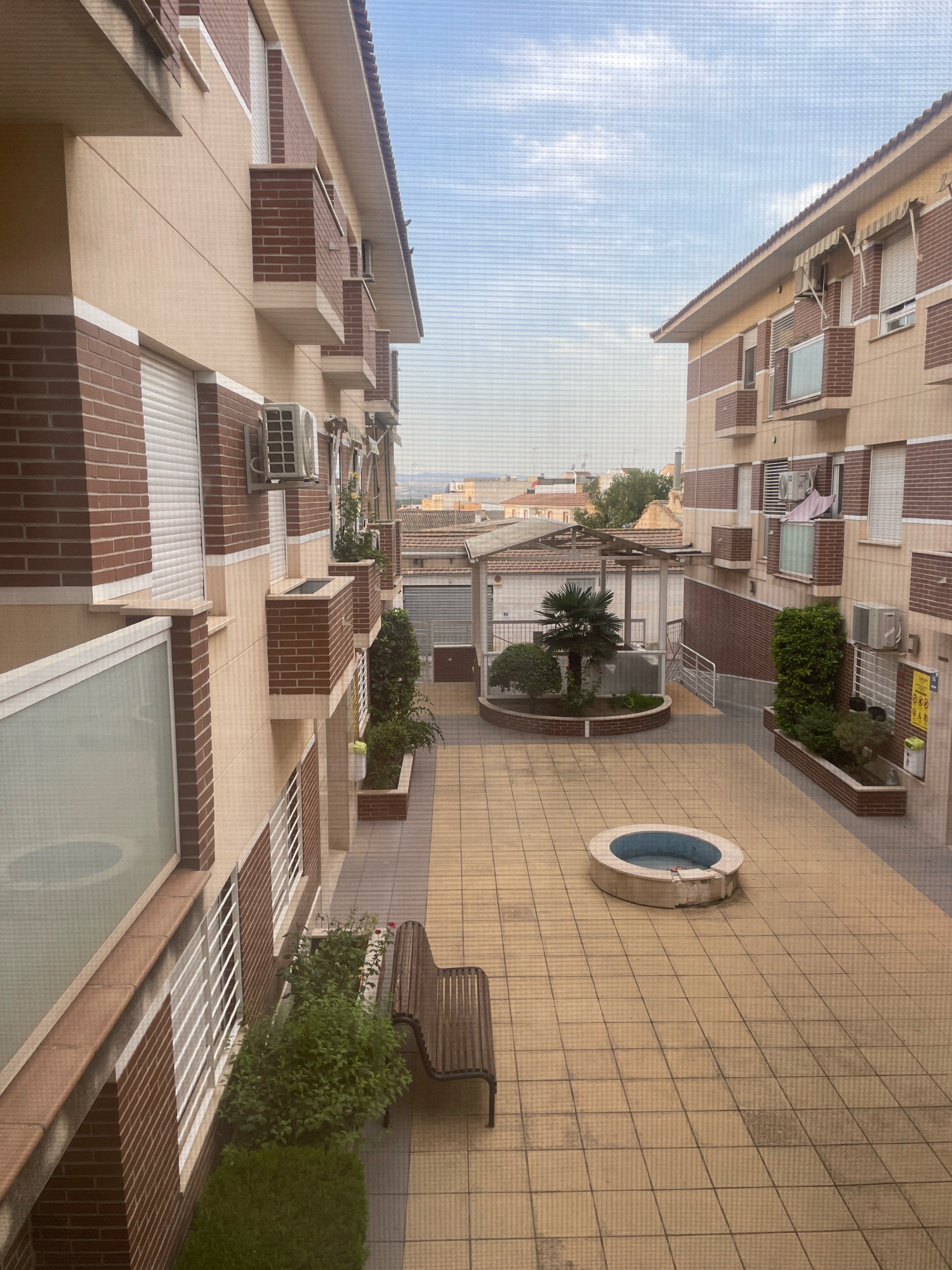 The image shows a courtyard between two apartment buildings, with a tiled floor and an oval-shaped fountain in the center. The buildings are constructed with light-colored stucco and red brick accents, featuring multiple balconies with white railings. Air conditioning units are visible on the exterior walls of the buildings. A wooden bench with a metal frame sits to the left of the courtyard, near some greenery and a small tree. In the background, there are more buildings, some with tiled roofs, and a distant view of the sky. The overall scene suggests a residential area in Atarfe, Spain.