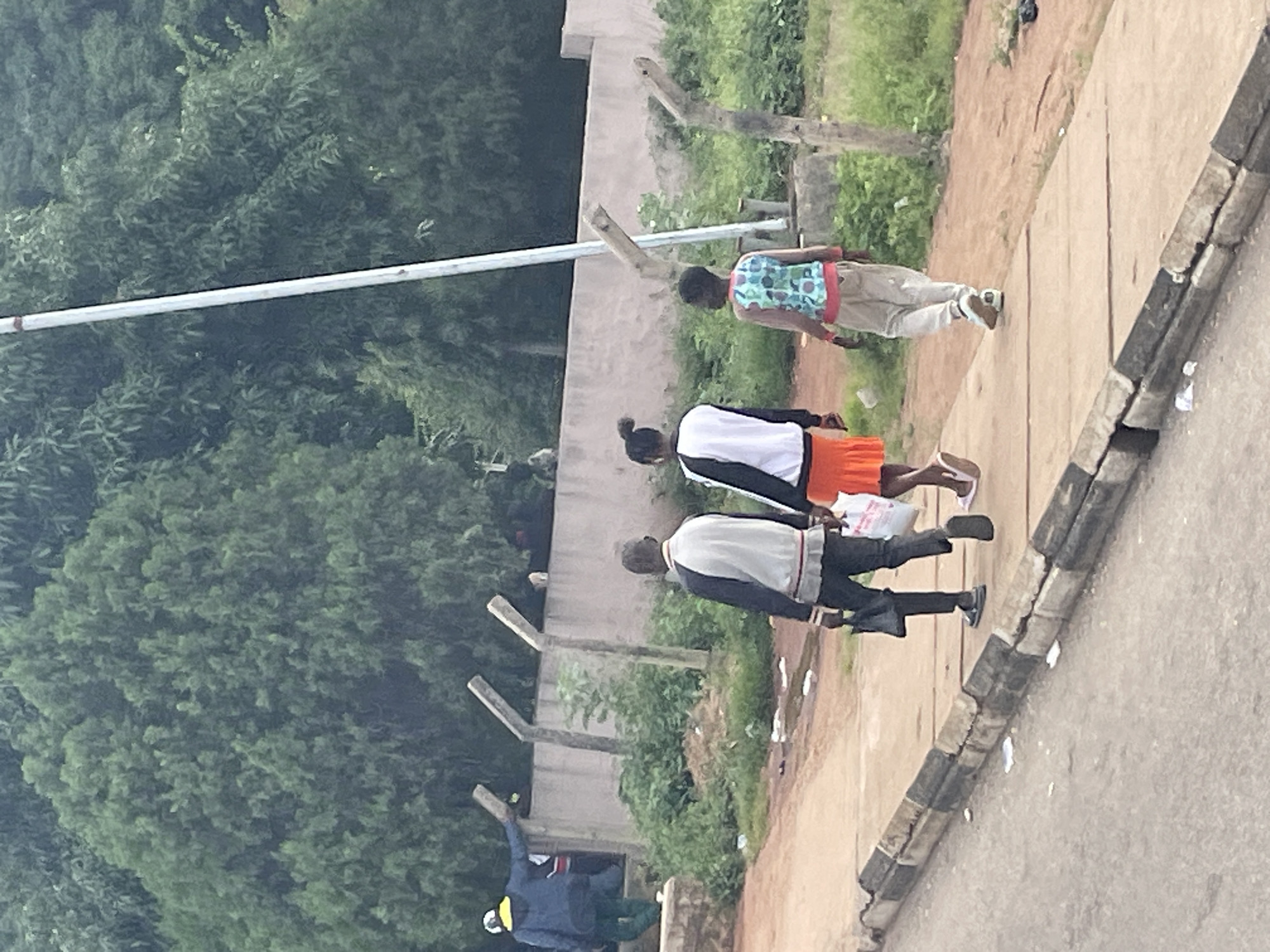 The image depicts a street scene in Jos, Nigeria, on what appears to be an overcast day. In the foreground, a paved sidewalk runs alongside a dirt road. The sidewalk is bordered by a concrete curb with alternating black and white painted sections, showing signs of wear and tear. Litter is scattered along the edge of the sidewalk and road.
Several people are walking on the sidewalk. From left to right, a person in a blue jacket with a yellow hood is seen walking away from the camera, appearing to ascend a small embankment. Further down the sidewalk, three individuals are walking towards the camera. The first is a man in a black and white jacket, carrying a black bag in his left hand. He is wearing dark pants and sandals. Next to him is a woman wearing a white jacket with black sleeves and an orange skirt. She is carrying a white plastic bag in her right hand. The third person is a young individual wearing a light-colored patterned top and light-colored pants, walking with his hands at his sides.
Behind the sidewalk is a low concrete wall with concrete posts extending upwards, suggesting a fence. Beyond the wall, dense green foliage and trees form a backdrop, indicating a somewhat natural or overgrown environment within or bordering the urban area. A tall, light-colored pole, possibly a utility pole, is visible amidst the trees. The overall impression is of an outdoor public space, likely a street or pathway in a residential or commercial area of Jos.