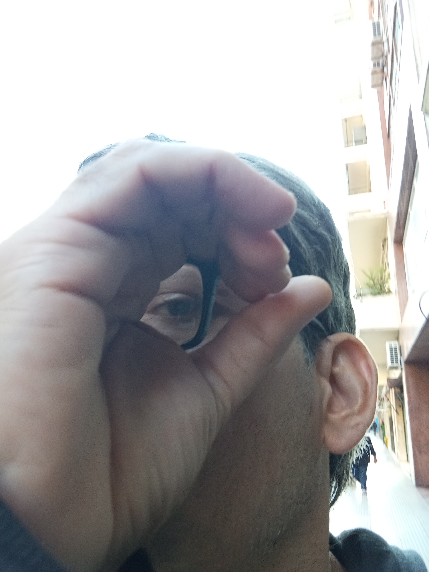 A close-up shot taken from a low angle depicts a person obscuring their face with their hand, which also holds a pair of eyeglasses. The person has short, dark gray hair and light skin. Part of an ear is visible. The person’s one visible eye looks directly at the camera through the eyeglasses.
In the background, a narrow street is flanked by tall buildings. One building is white with balconies. An air conditioning unit is visible on one balcony. On the sidewalk, at least one person is walking away from the camera. The image was taken outdoors during daylight.
The presence of balconies, architectural style, and air conditioning units are consistent with a city environment like Buenos Aires, Argentina.